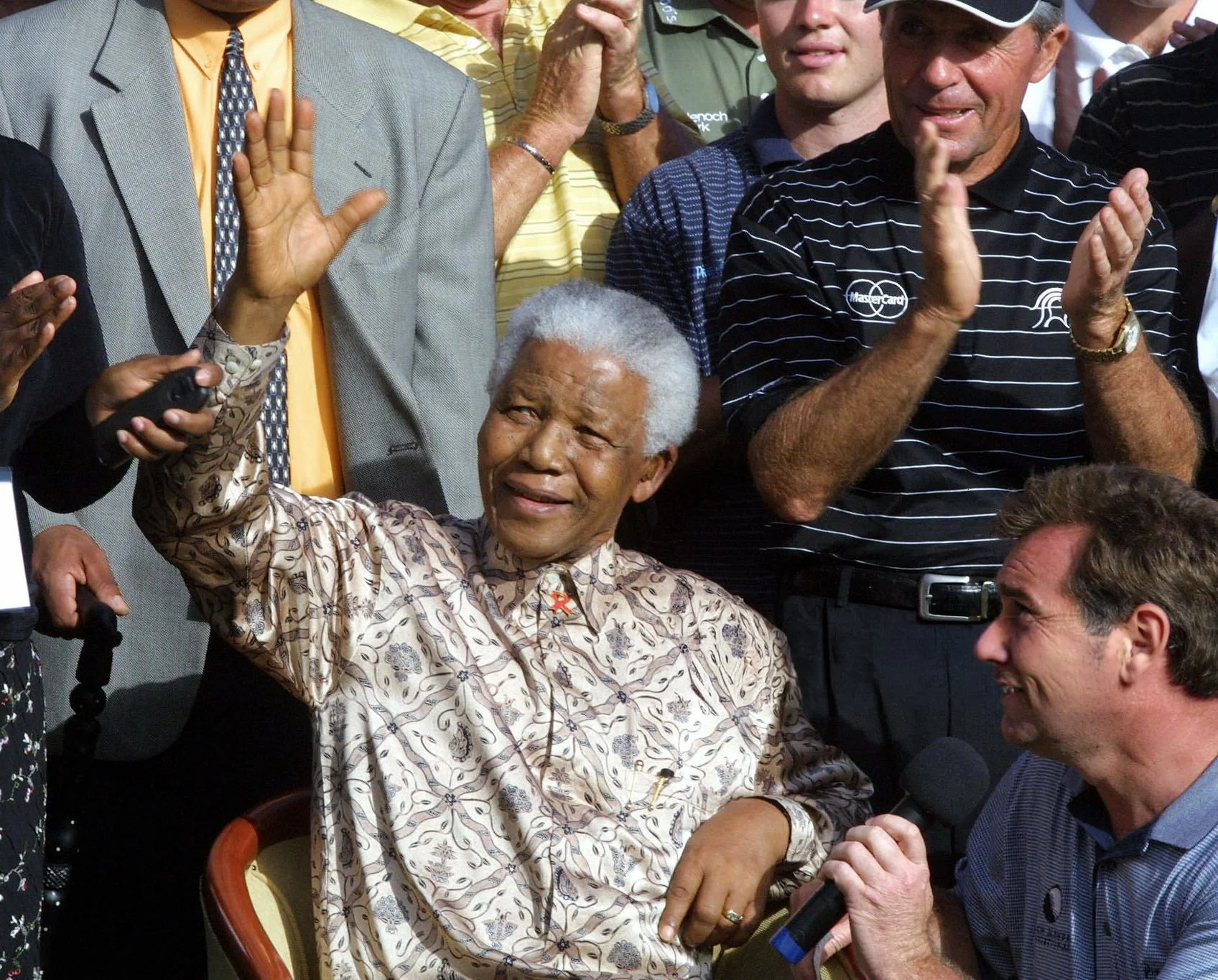 A group of people gathered around an elderly man with gray hair, who is seated and waving. The elderly man is wearing a patterned shirt, and several people are clapping and talking to him, including a man with a microphone and a man in a gray suit.