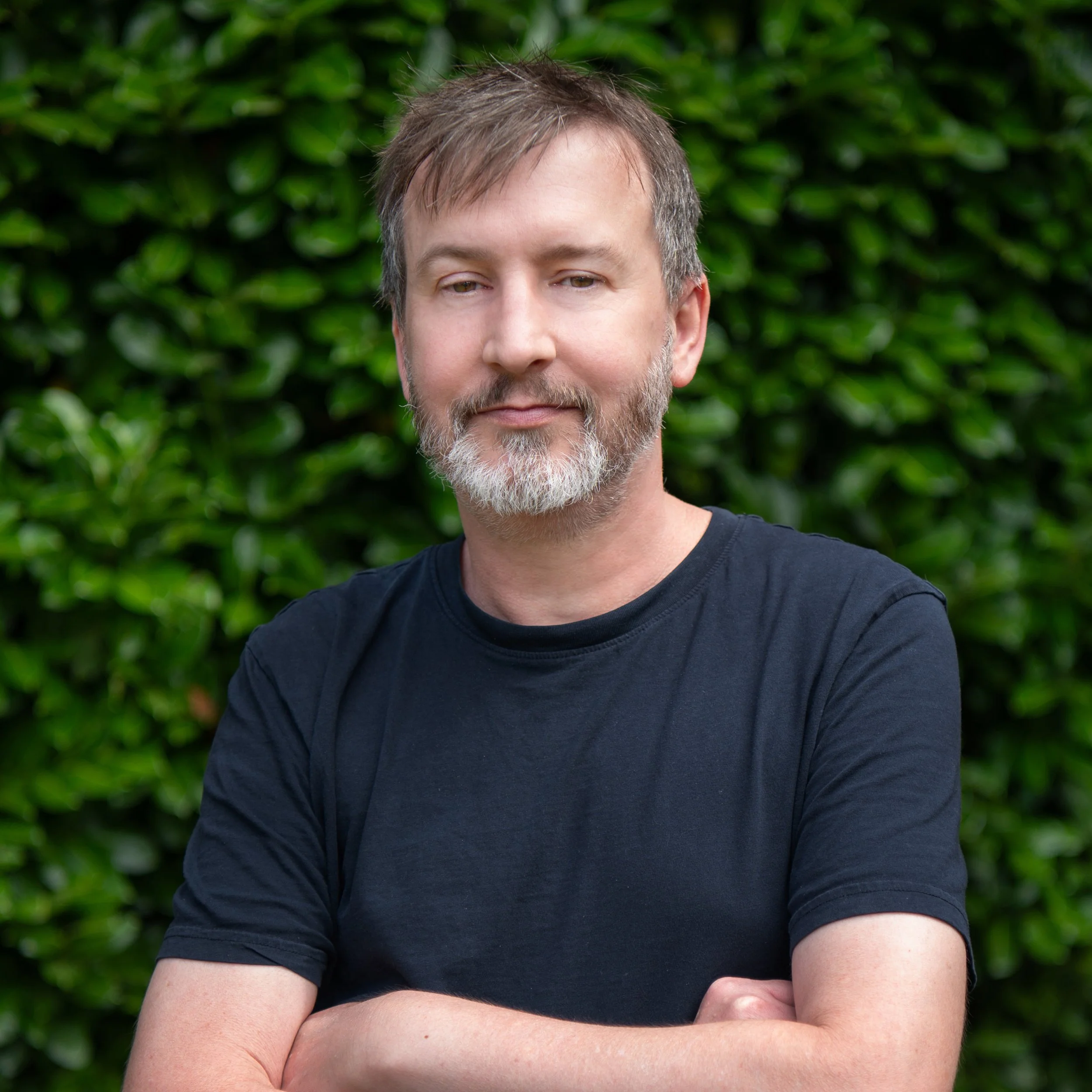 A man with gray hair and beard, wearing a black shirt, standing outdoors in front of green bushes.