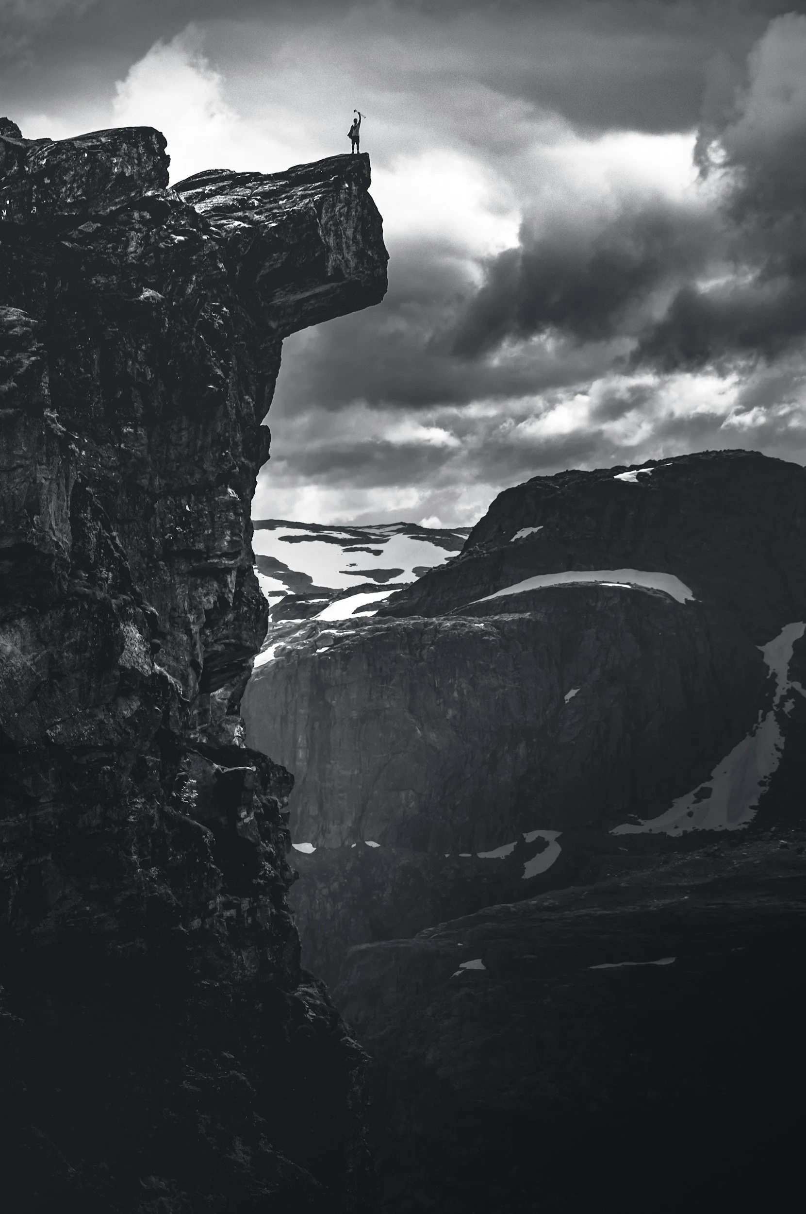 A person standing on a small rocky ledge at the top of a tall cliff, holding a stick or staff, overlooking a rugged mountainous landscape with snow patches and dramatic cloudy sky.