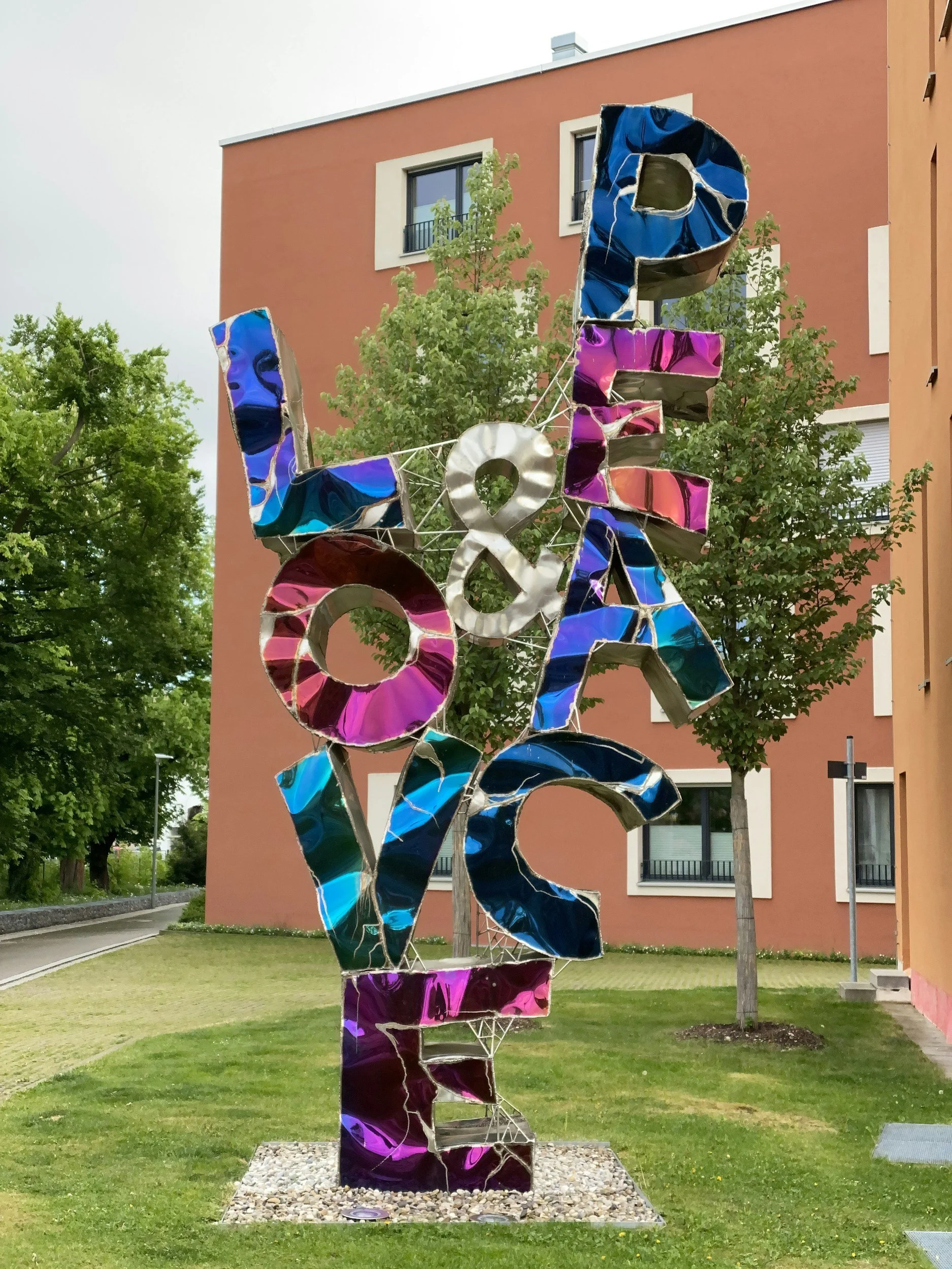 Colorful outdoor art installation spelling out 'LOVE' with a glittery ampersand in the middle, set on a small gravel base in front of green trees and a pastel orange building.