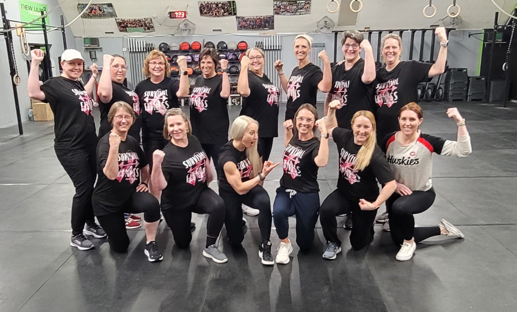 Group of women at a gym flexing their muscles, wearing matching black t-shirts with 'Survival Fitness' written on them, three of them kneeling in front and the rest standing behind, with gym equipment and a wall of helmets in the background.