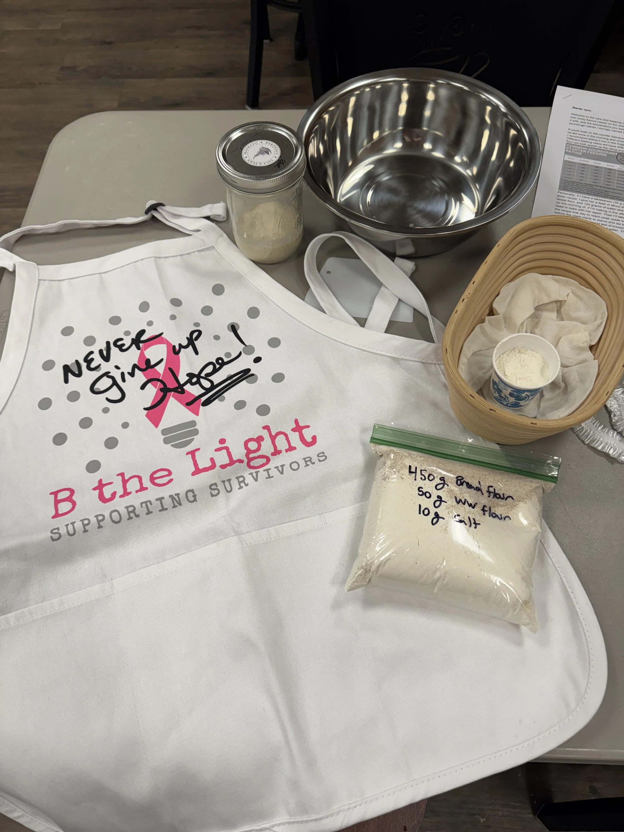 A table with a white apron supporting the text 'Never Give Up 2 the Light,' a glass jar of flour, a metal mixing bowl, and a small container of flour inside a woven basket, with a piece of paper with recipes or instructions in the background.