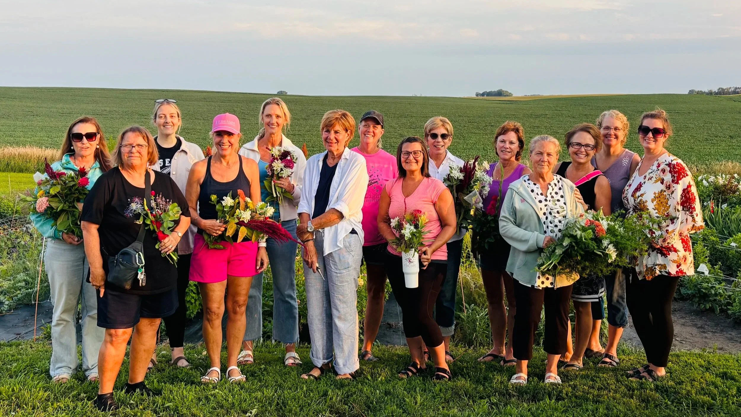A large group of women standing outdoors in a field, holding flowers, with green fields and a cloudy sky in the background.