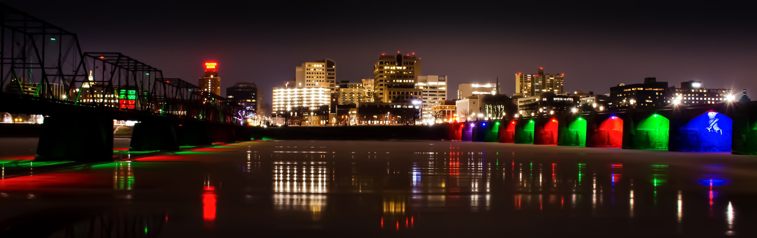 Nighttime cityscape with illuminated buildings and a bridge decorated with colorful lights reflected on the water below.
