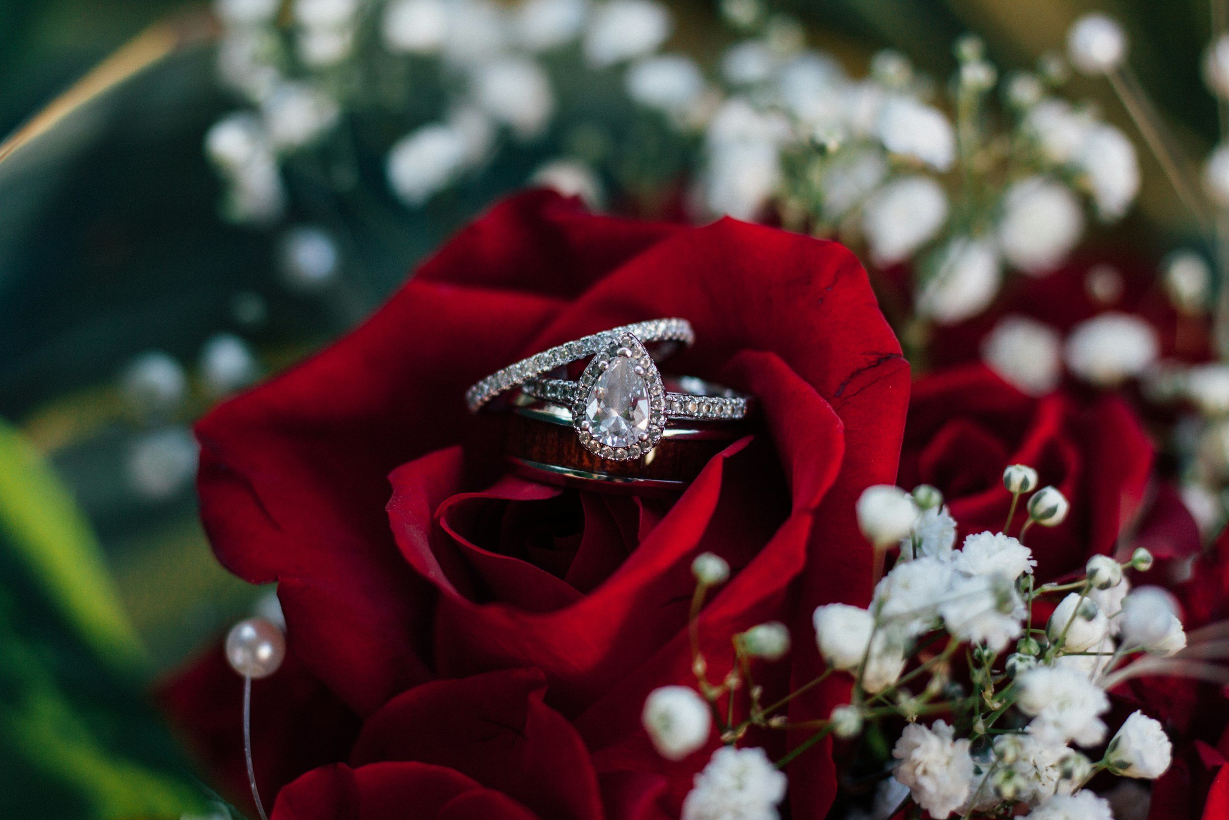 A close-up of a wedding ring set with a pear-shaped diamond, placed on a red rose surrounded by small white flowers and pearl accents.