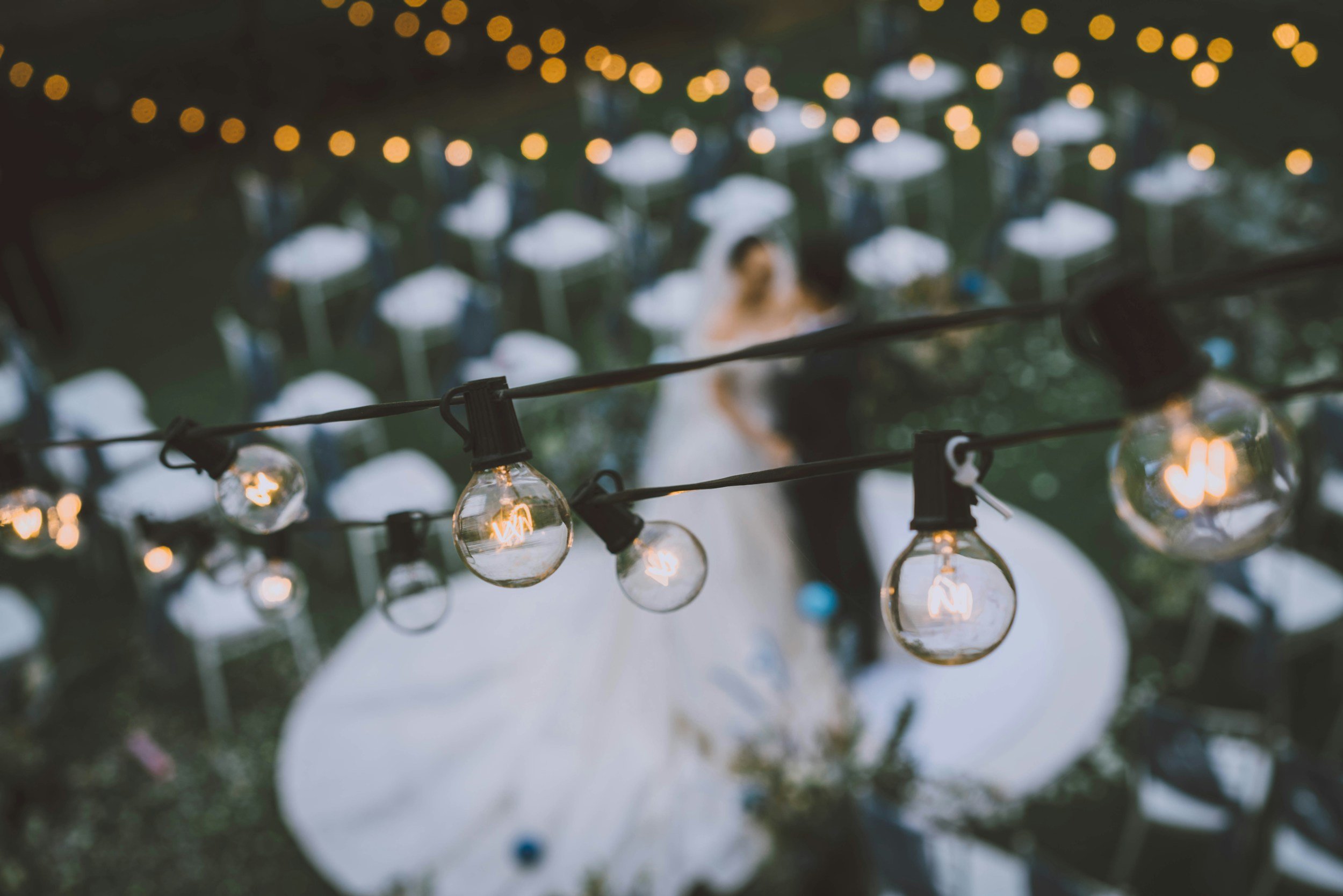 String of glowing light bulbs hanging outdoors at a wedding reception, with a blurred bride and groom in the background and rows of empty chairs.
