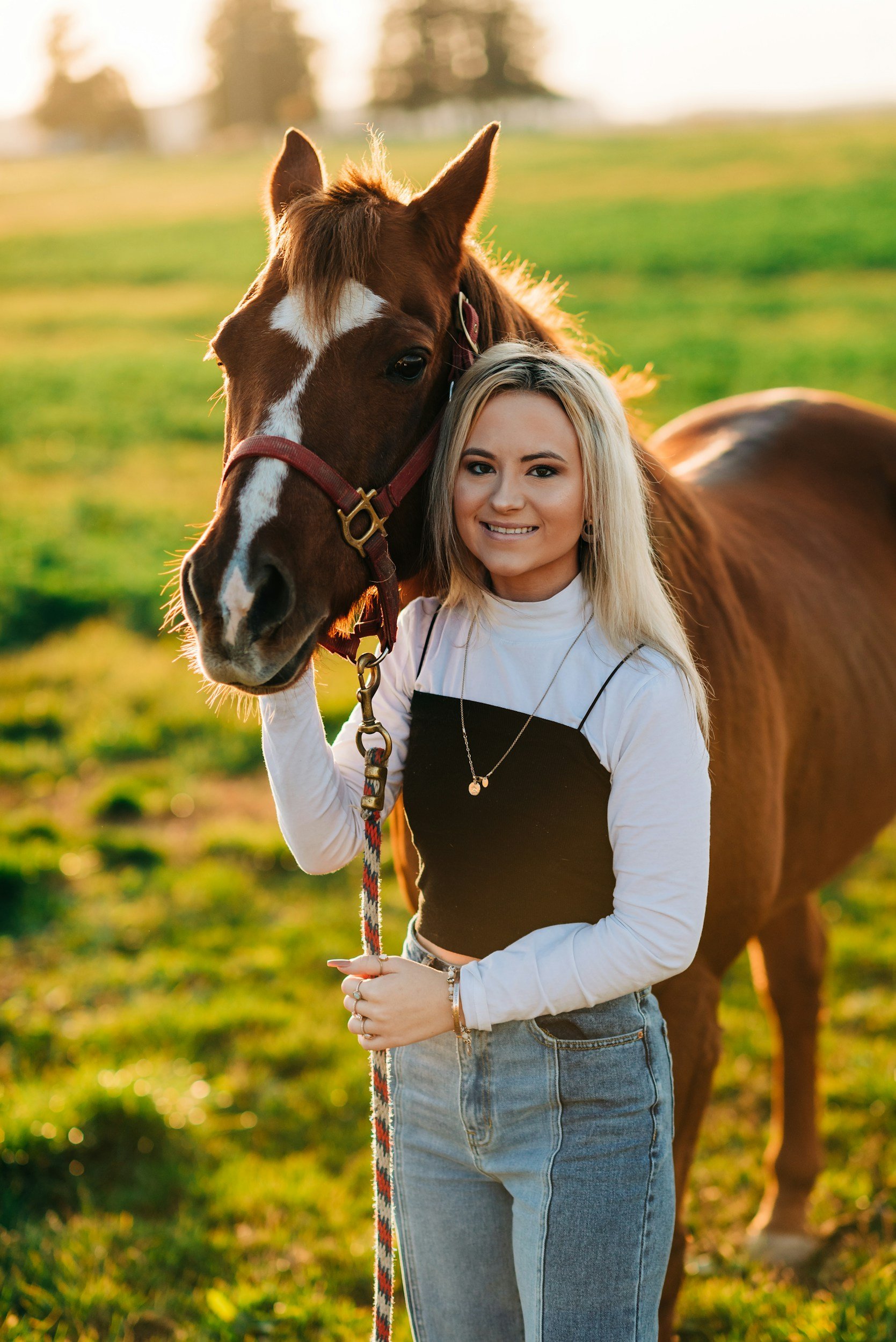 A young woman with blonde hair smiling and holding a brown horse by the reins in a green field during sunset.
