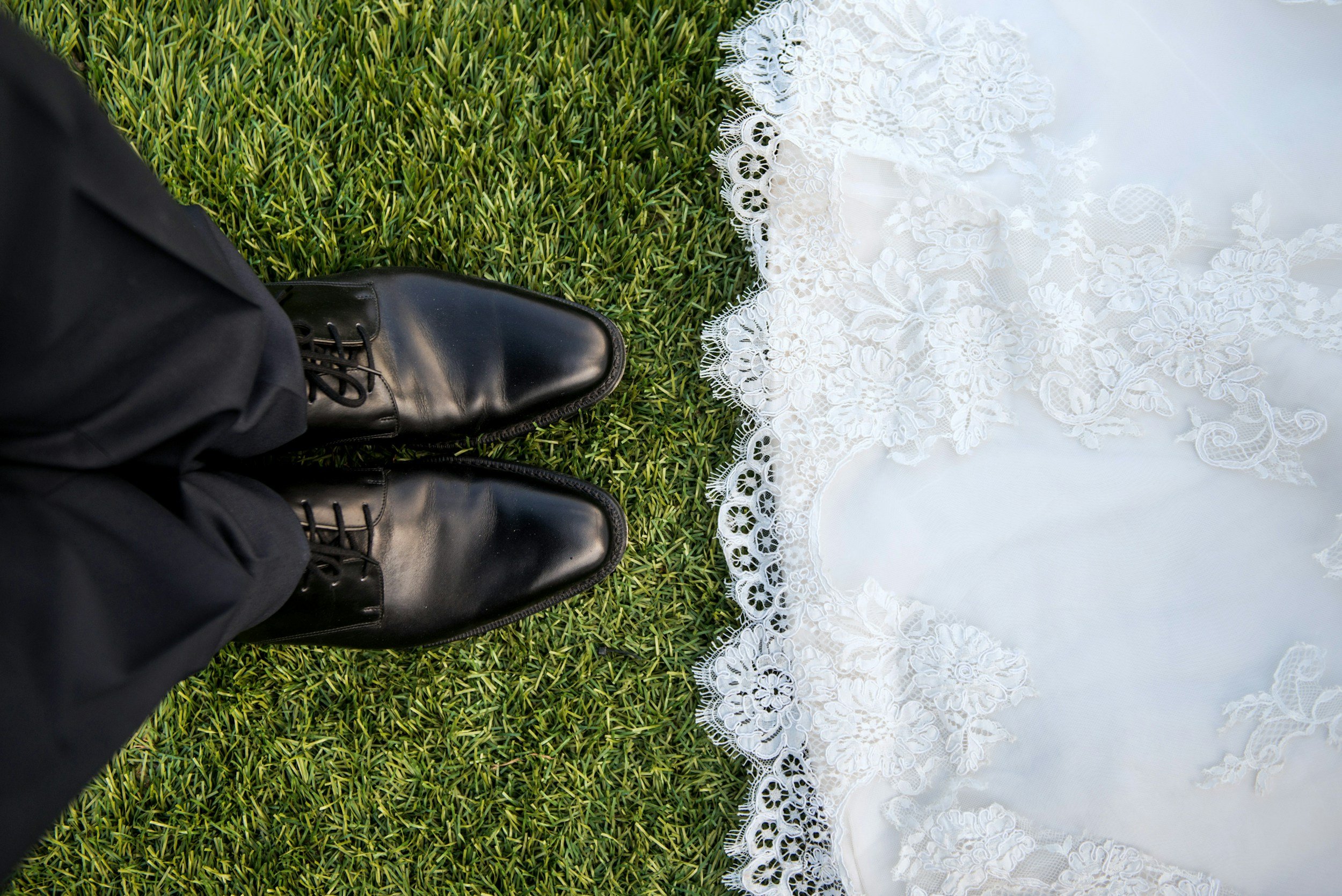 Black polished dress shoes and black pants on green grass next to a white lace wedding dress.