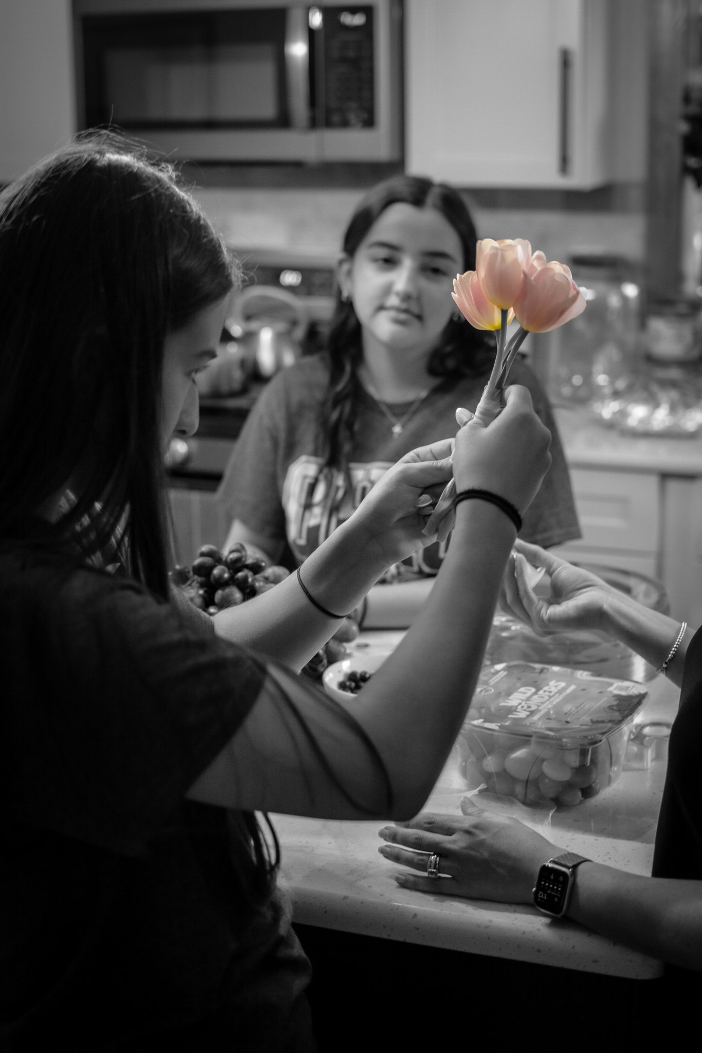 A girl holding a bouquet of pink tulips, with a woman pointing at the flowers, while another girl looks on, in a kitchen setting.