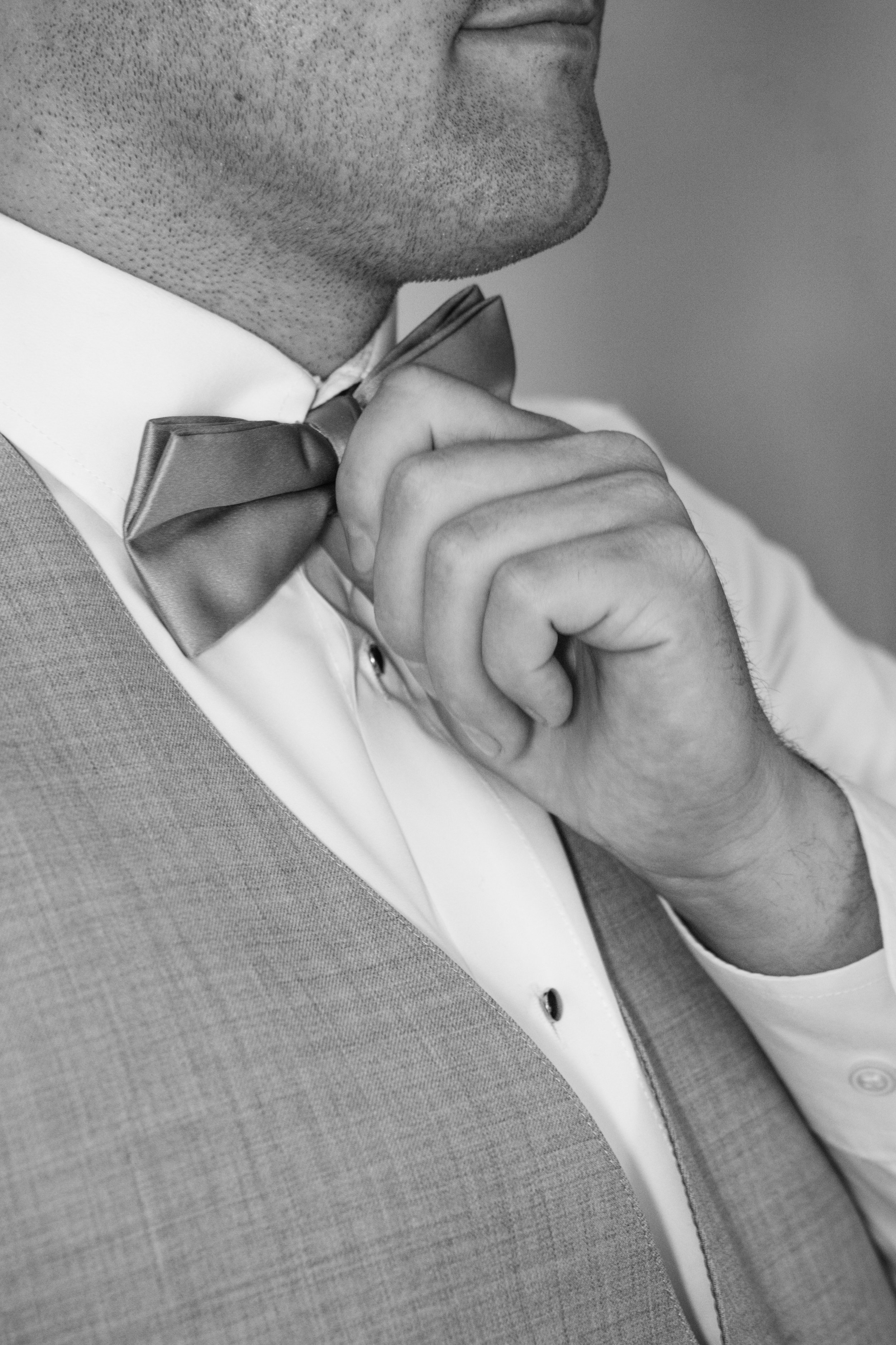 Close-up of a man adjusting a bow tie, wearing a white dress shirt, a gray vest, and a bow tie.