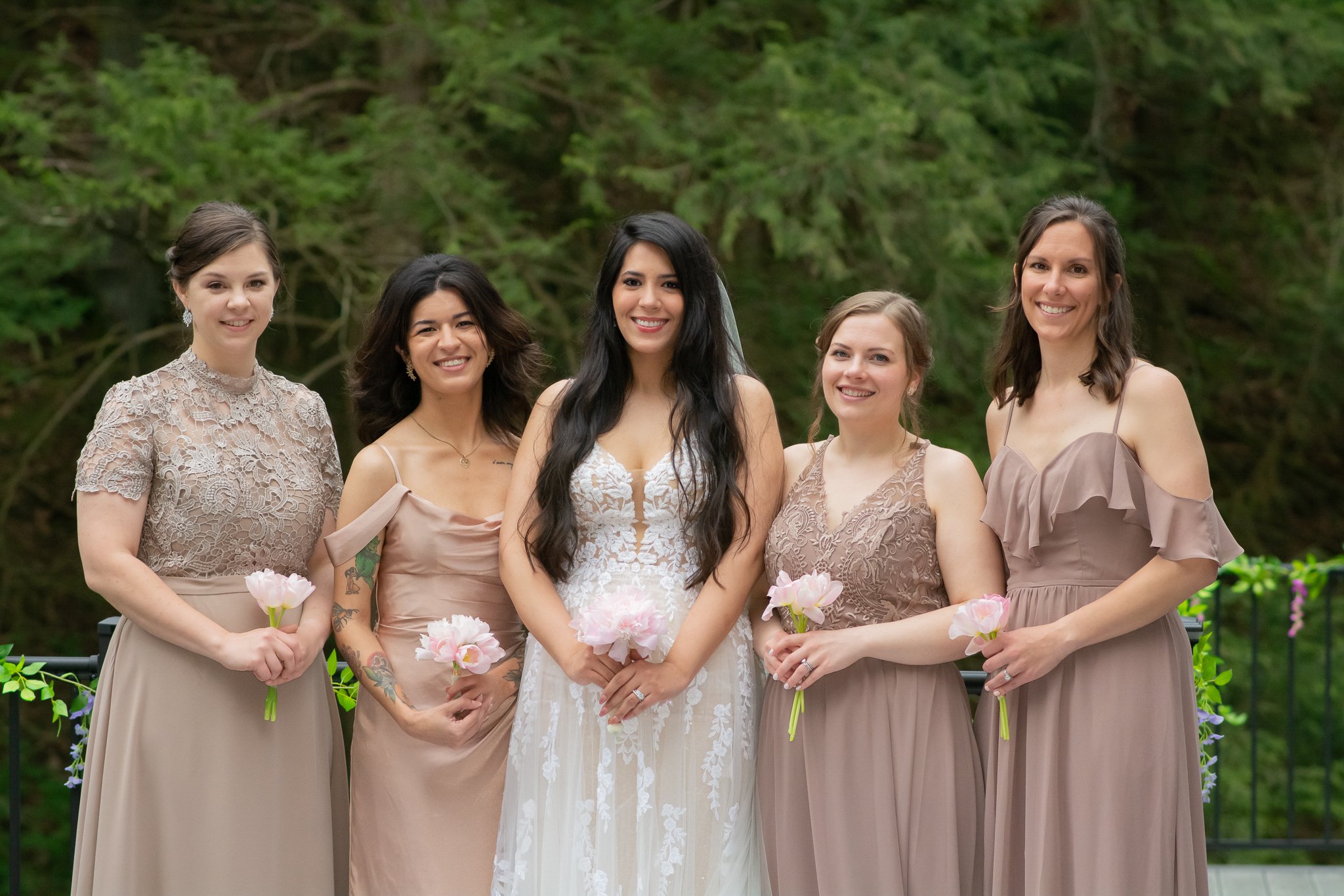 A bride in a white lace wedding dress standing with five bridesmaids in beige dresses, smiling and holding pink flowers outdoors with green foliage in the background.
