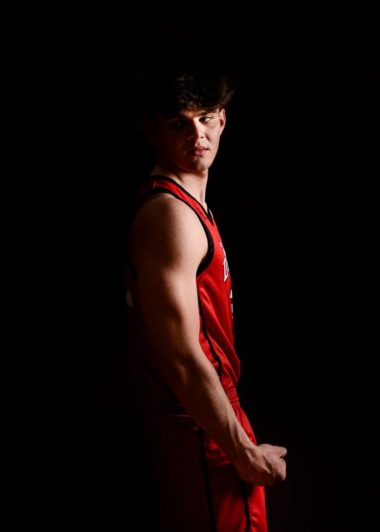 Young male basketball player in a red uniform, standing sideways with a serious expression against a black background.
