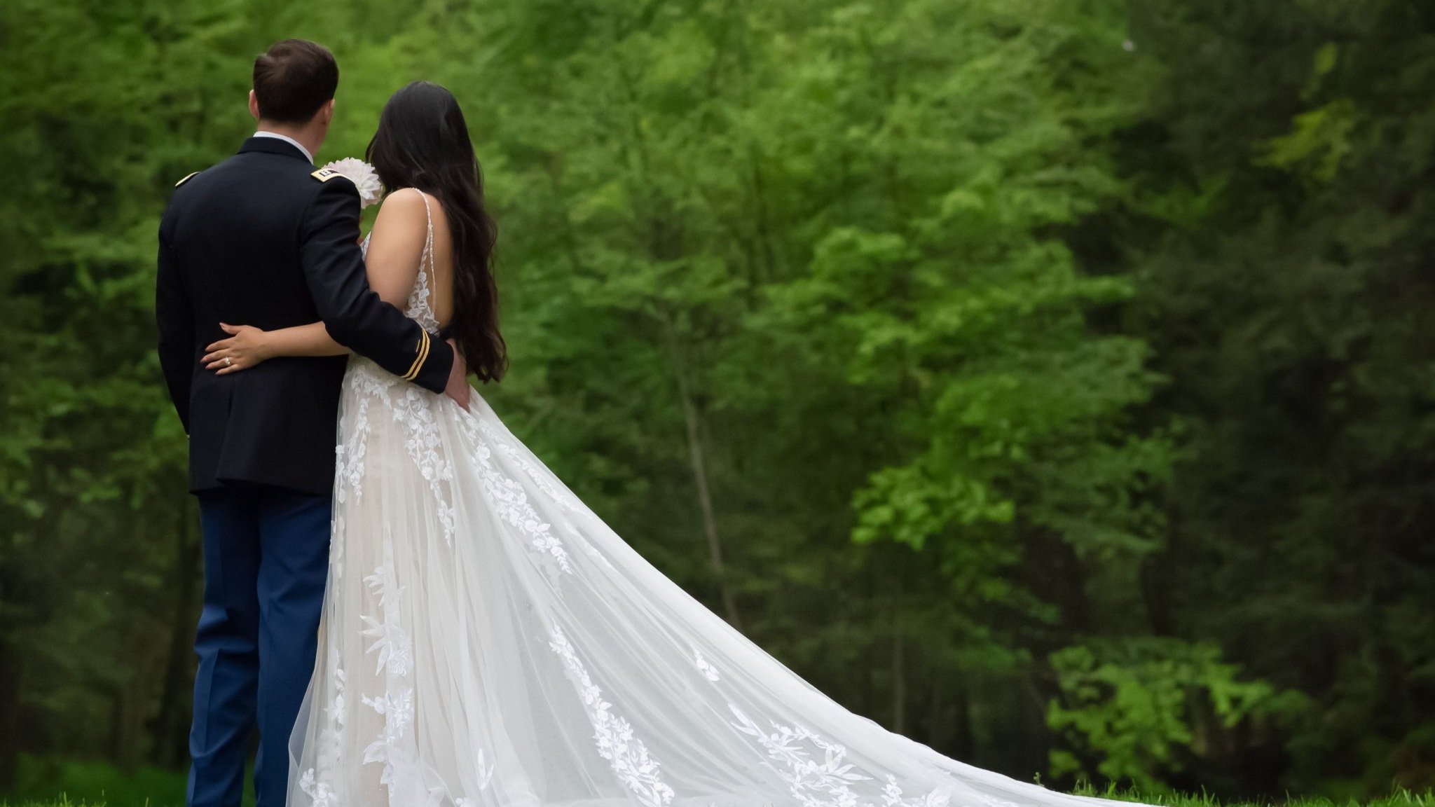 A bride and groom standing together outdoors in a lush green forest, embracing each other during their wedding photoshoot.