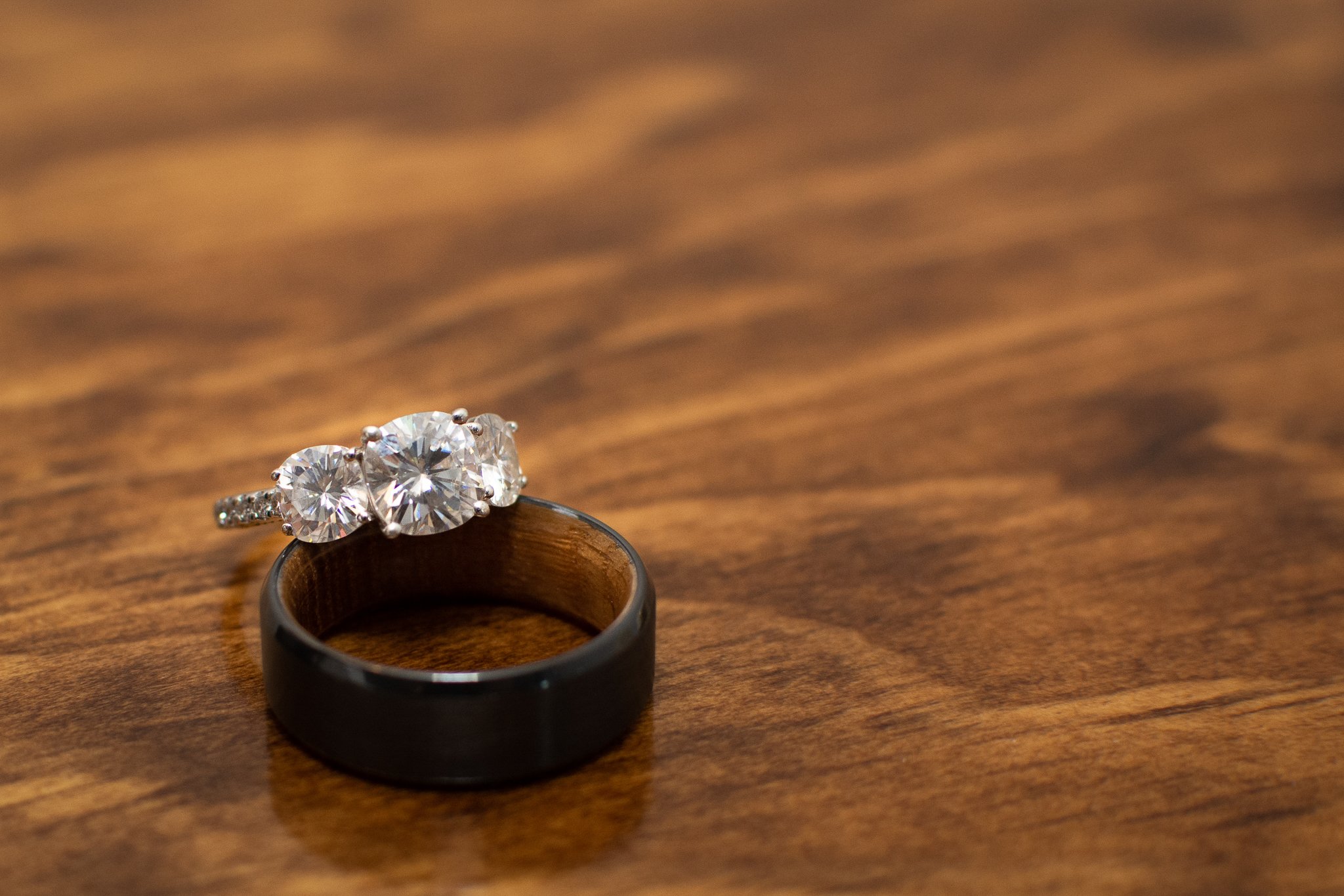 A black wedding band and an engagement ring with three large diamonds and smaller stones on a wooden surface.