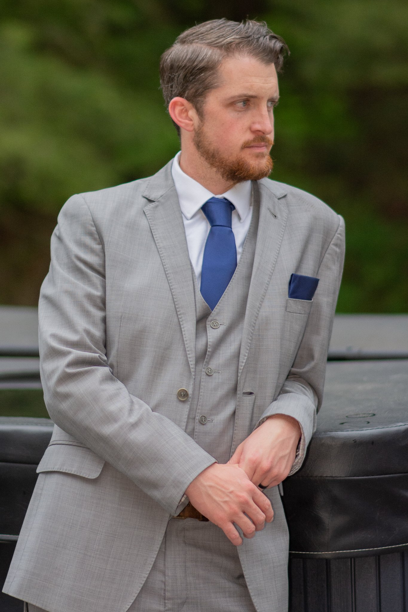 A man in a light gray suit with a blue tie and pocket square standing outdoors near a black trash bin.