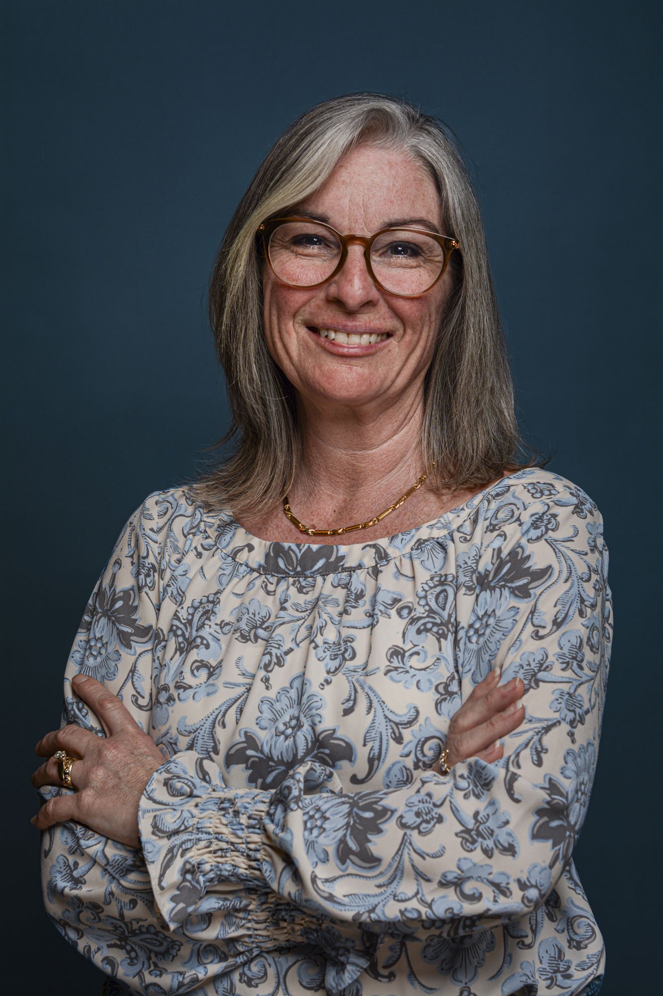 Middle-aged woman with gray hair, glasses, smiling with arms crossed, wearing a floral blouse and gold jewelry, against a dark blue background.