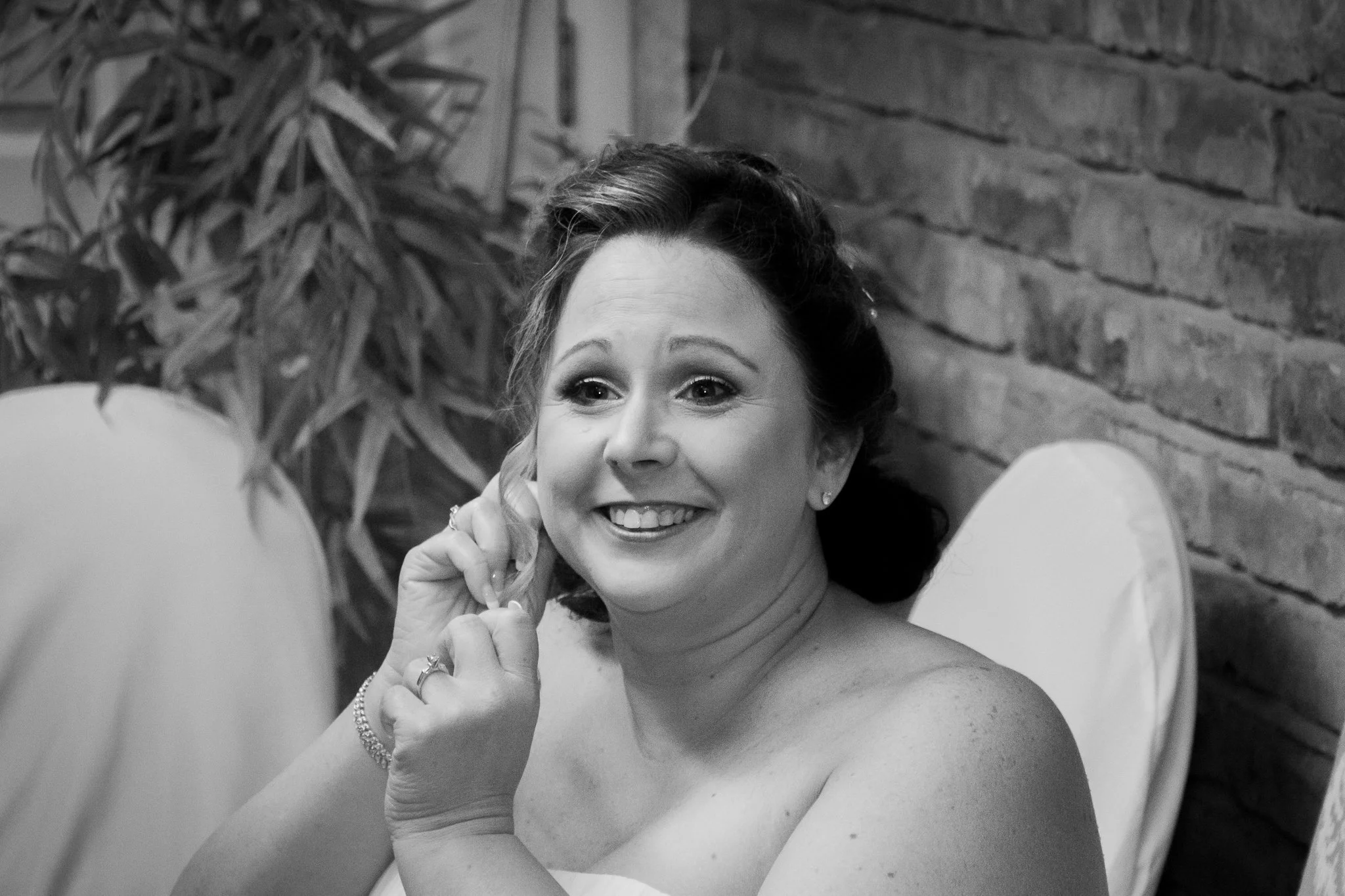 A smiling woman with dark hair styled in an elegant updo, wearing earrings and a strapless dress, sitting in front of a brick wall and a potted plant.