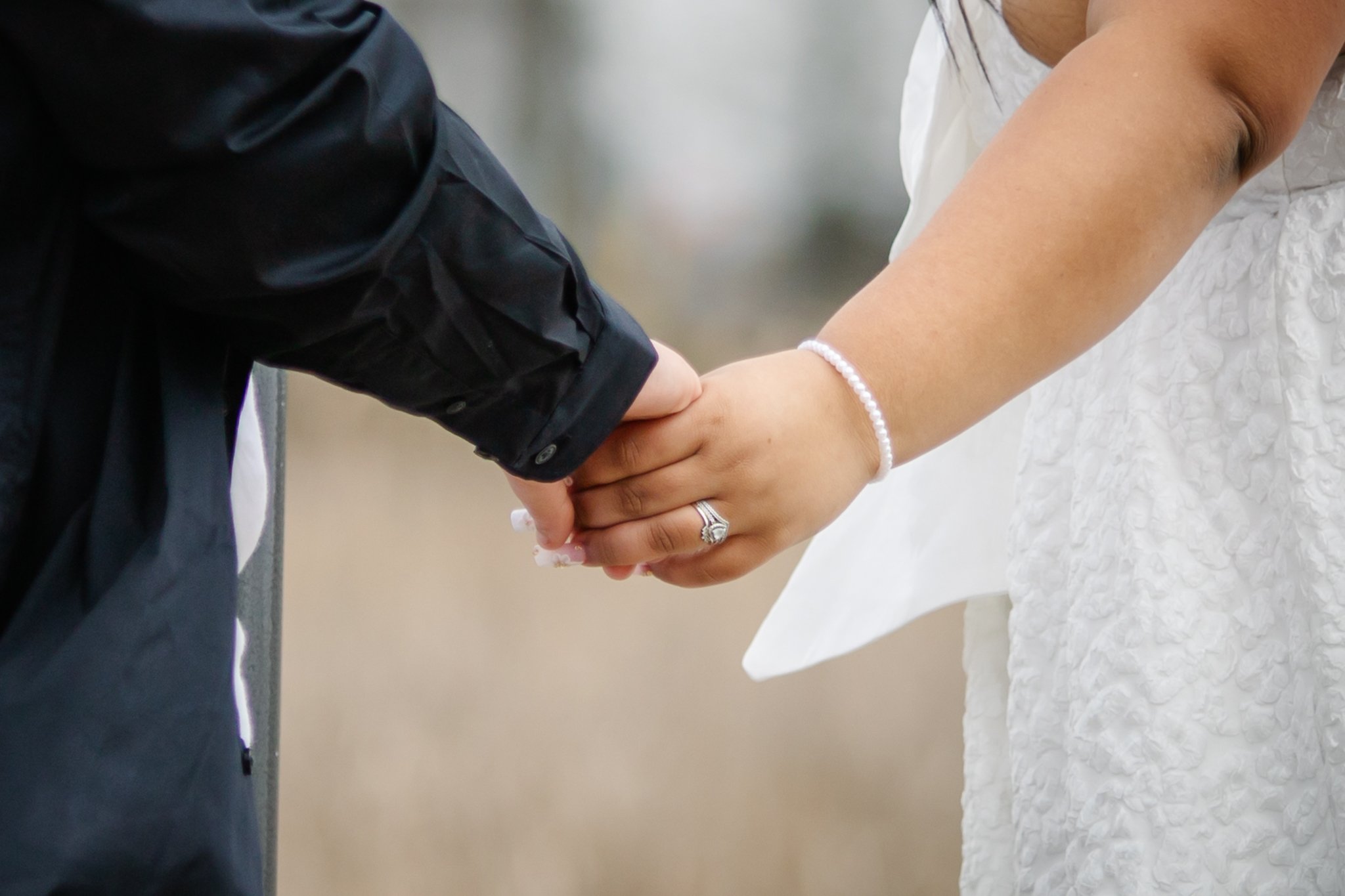 Close-up of a couple holding hands during a wedding ceremony, with the bride wearing a white dress and a pearl bracelet, and the groom in a black jacket.