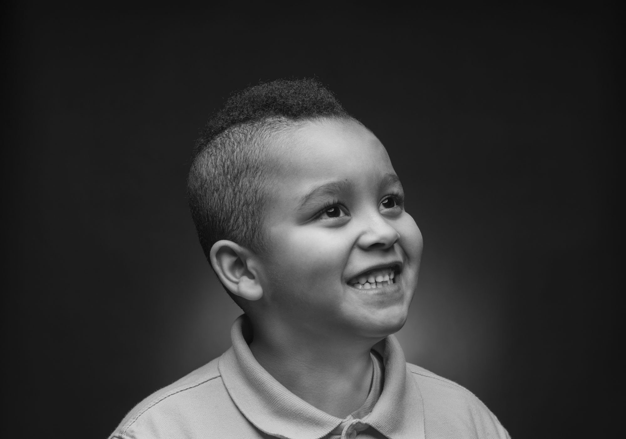 Black and white portrait of a young boy with a wide smile, wearing a collared shirt, looking upwards with a joyful expression.