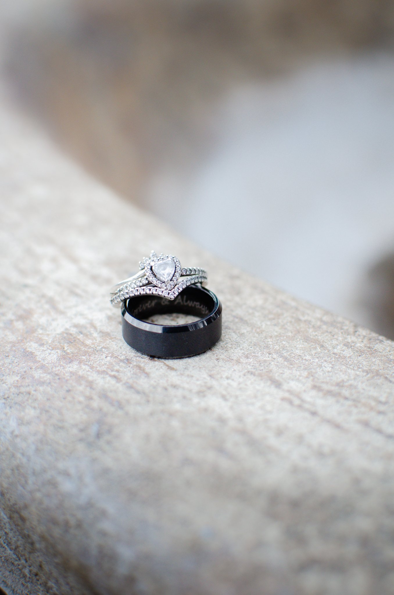 A black wedding band and a silver engagement ring with a heart-shaped diamond, both resting on a textured stone surface.