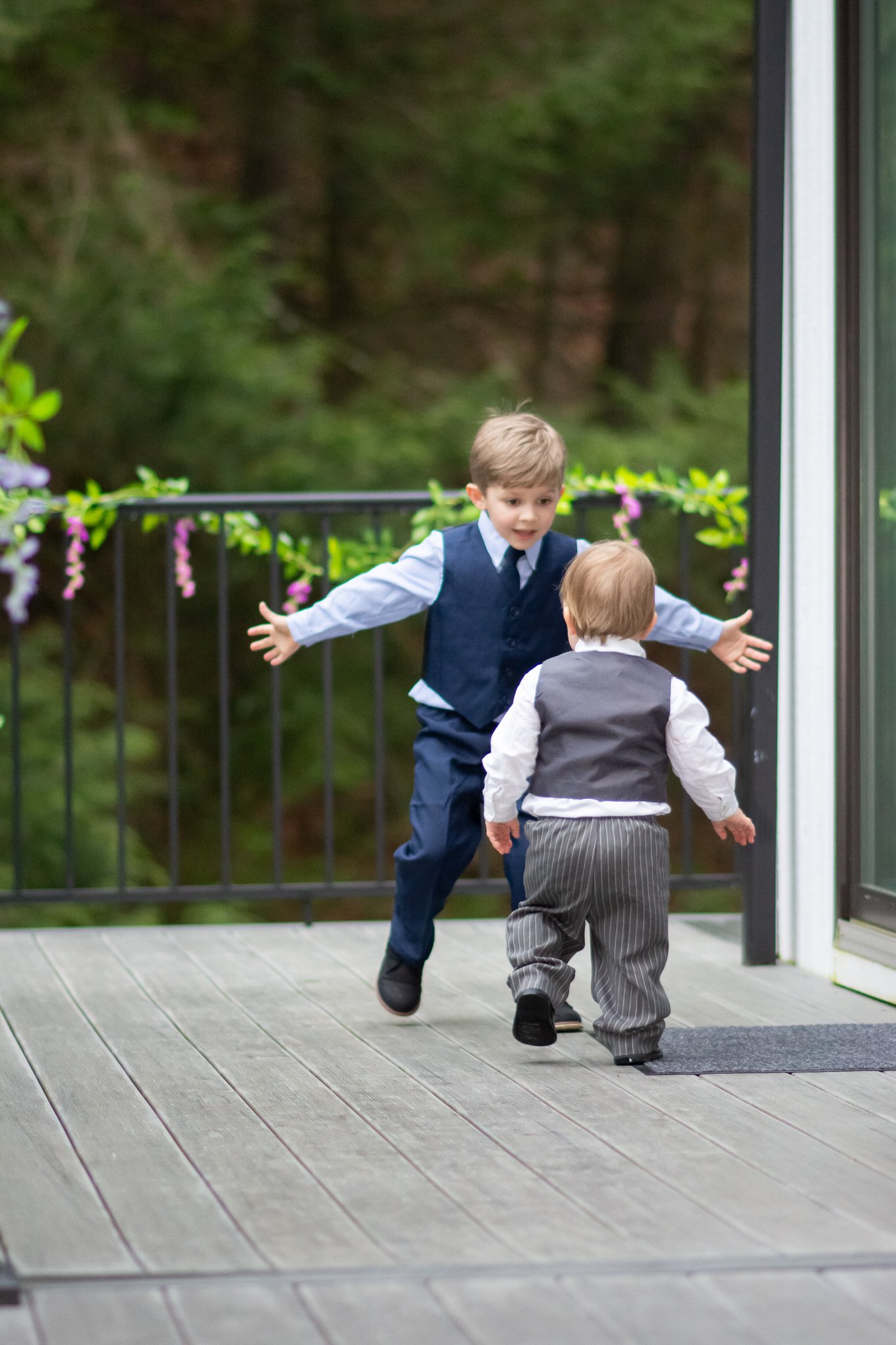 Two young boys in formal attire playing on a wooden deck outdoors near a sliding glass door, with green trees and purple flowers in the background.