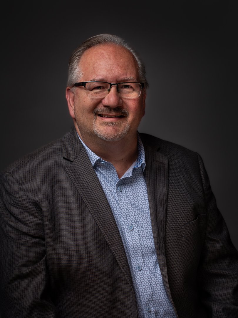 Portrait of a smiling middle-aged man with glasses, wearing a dark checked blazer and a patterned light blue button-up shirt, against a dark background.
