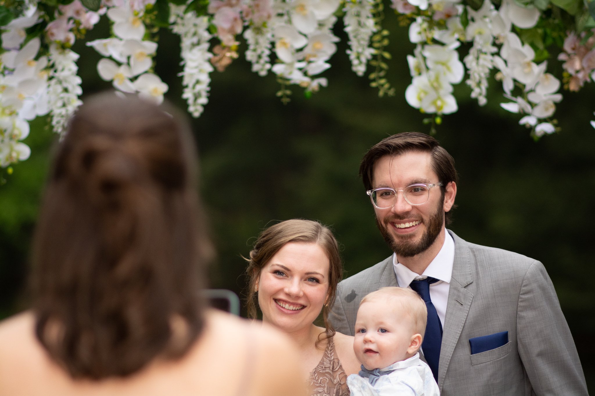 A happy family of three, including a woman, a man in a suit, and a young boy, taking a photo together at a wedding ceremony under an arch decorated with white flowers.