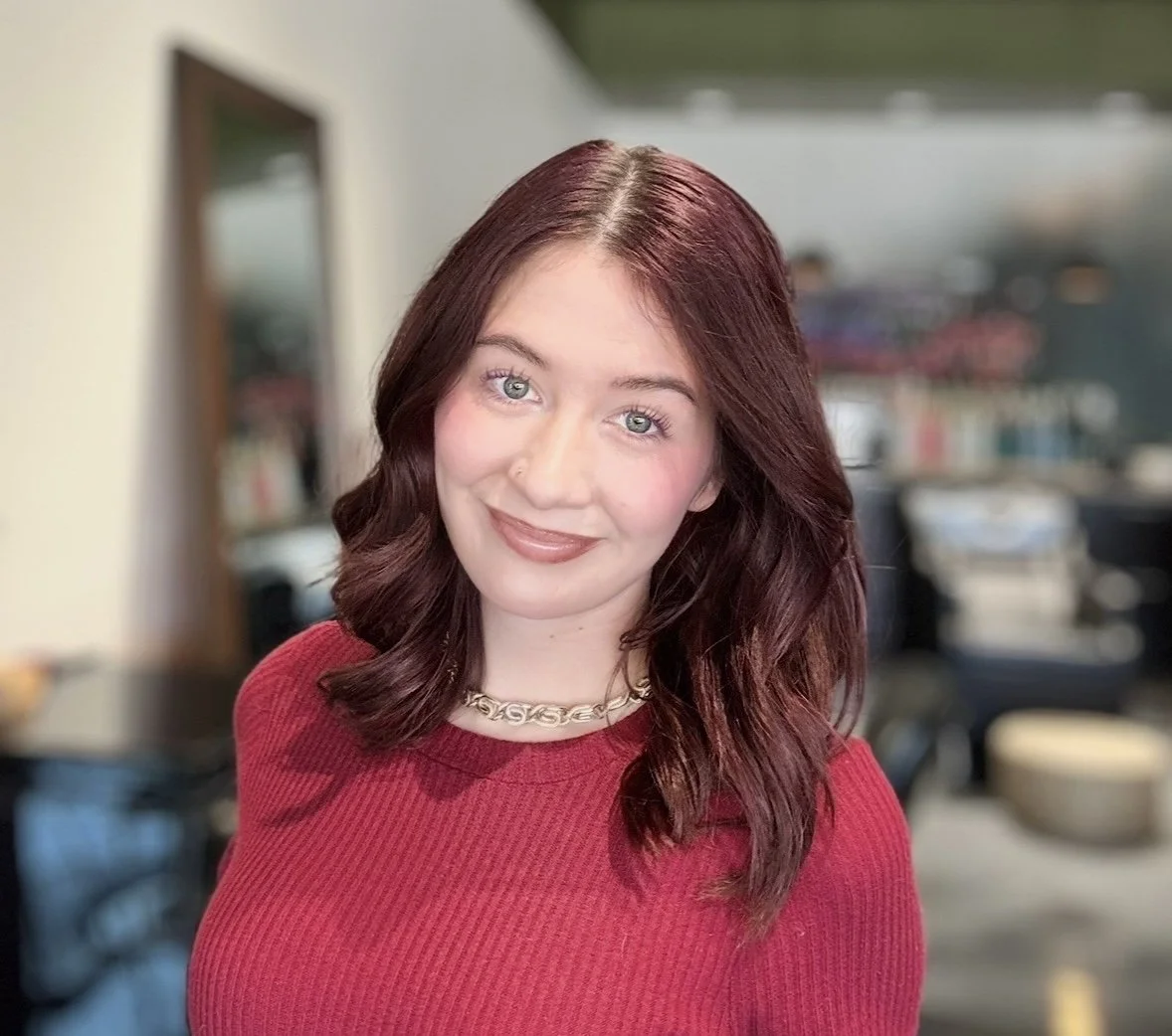 A woman with wavy, shoulder-length reddish-brown hair, wearing a red sweater and a gold chain necklace, smiling indoors with a blurred background.