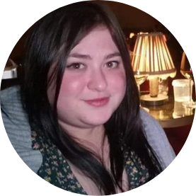 A young woman with long dark hair and fair skin, smiling slightly, sitting in a dimly lit setting with a table lamp in the background.