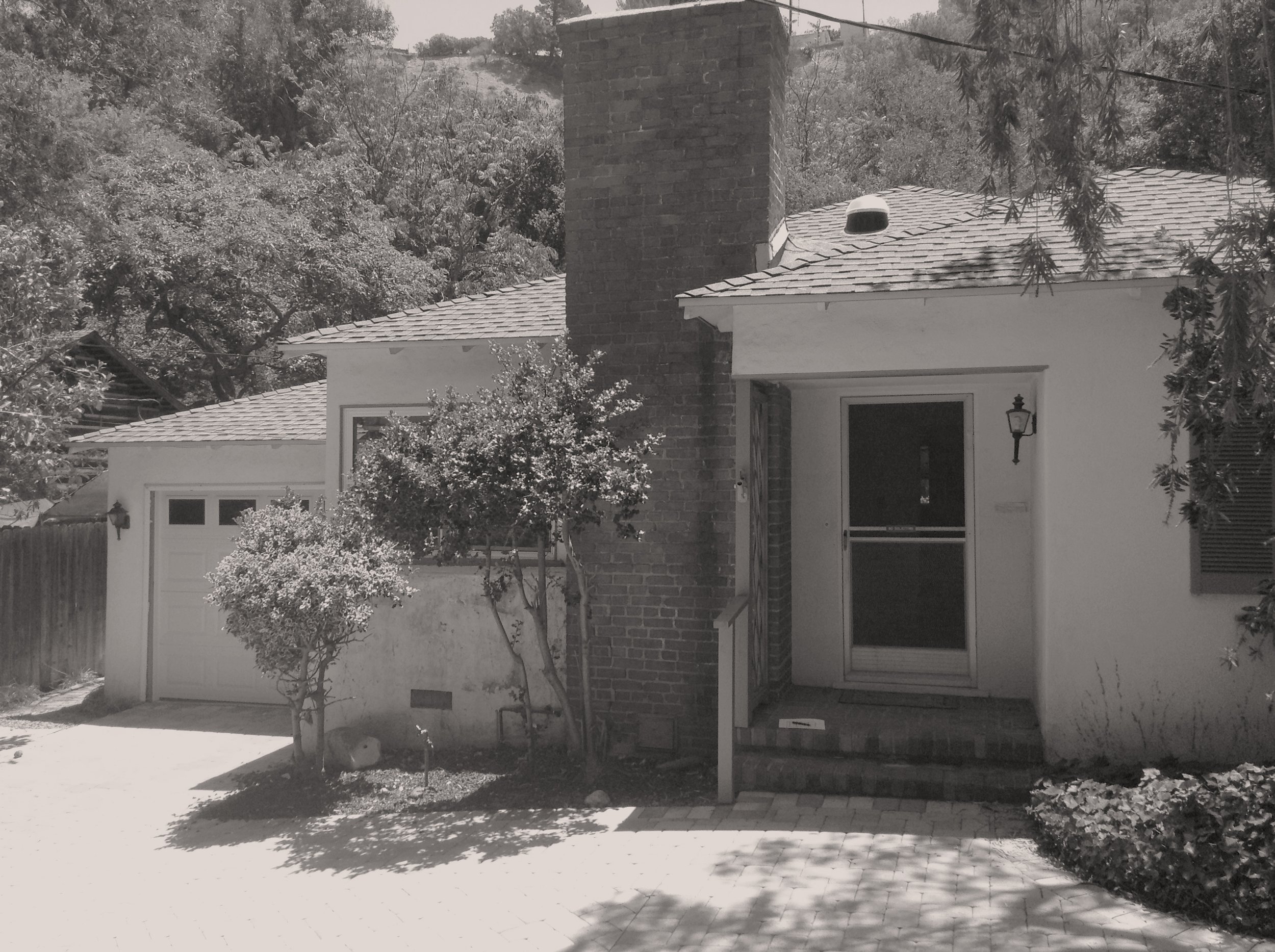 The image shows the front of a house with a brick chimney, a porch with steps, a screen door, and a lantern-style light fixture. There are two small trees and some bushes in the yard, with a paved walkway leading to the porch.