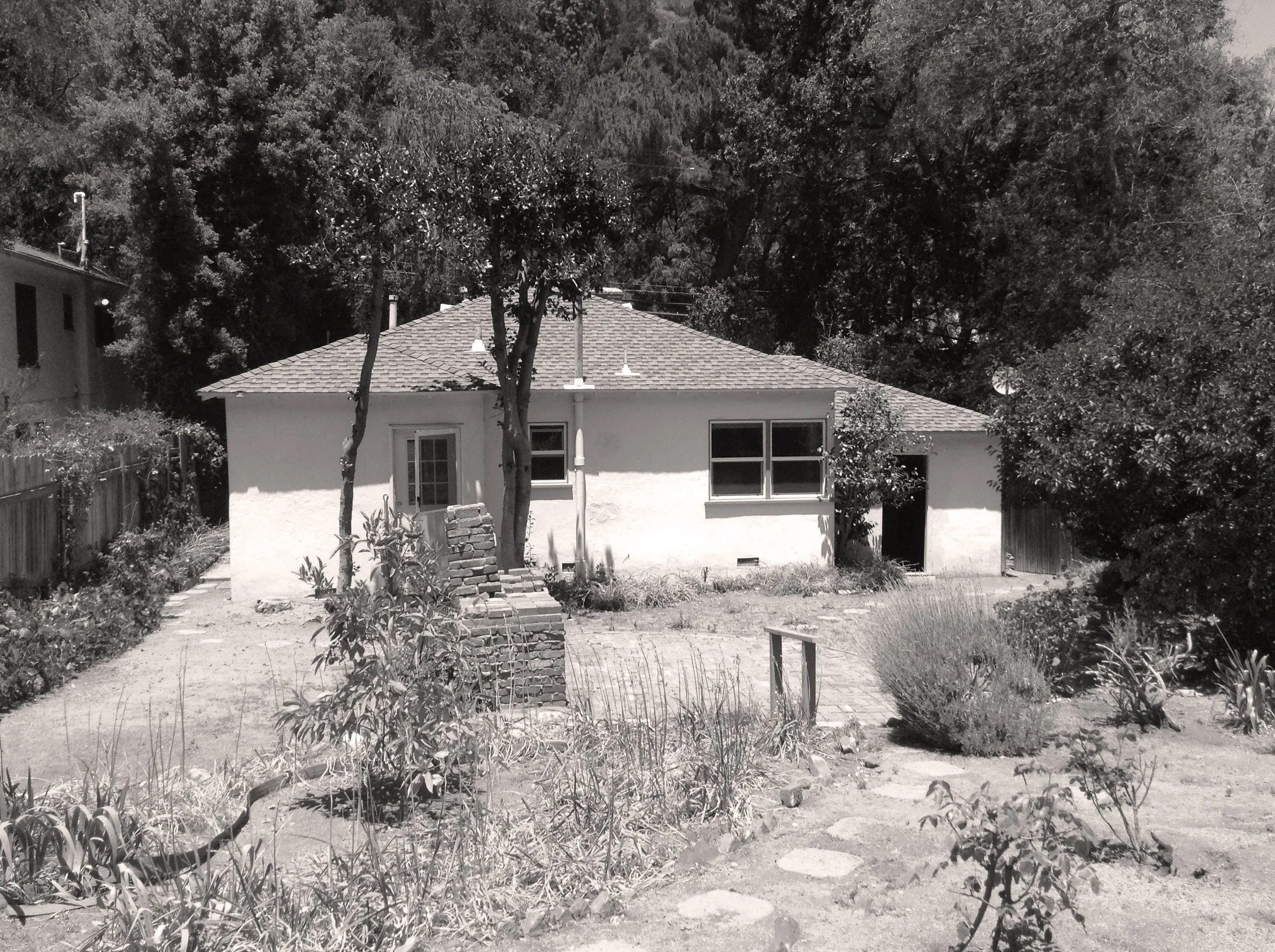 Black and white photo of a small, single-story house with a tiled roof, surrounded by trees and a garden with plants and a pathway leading to the house.