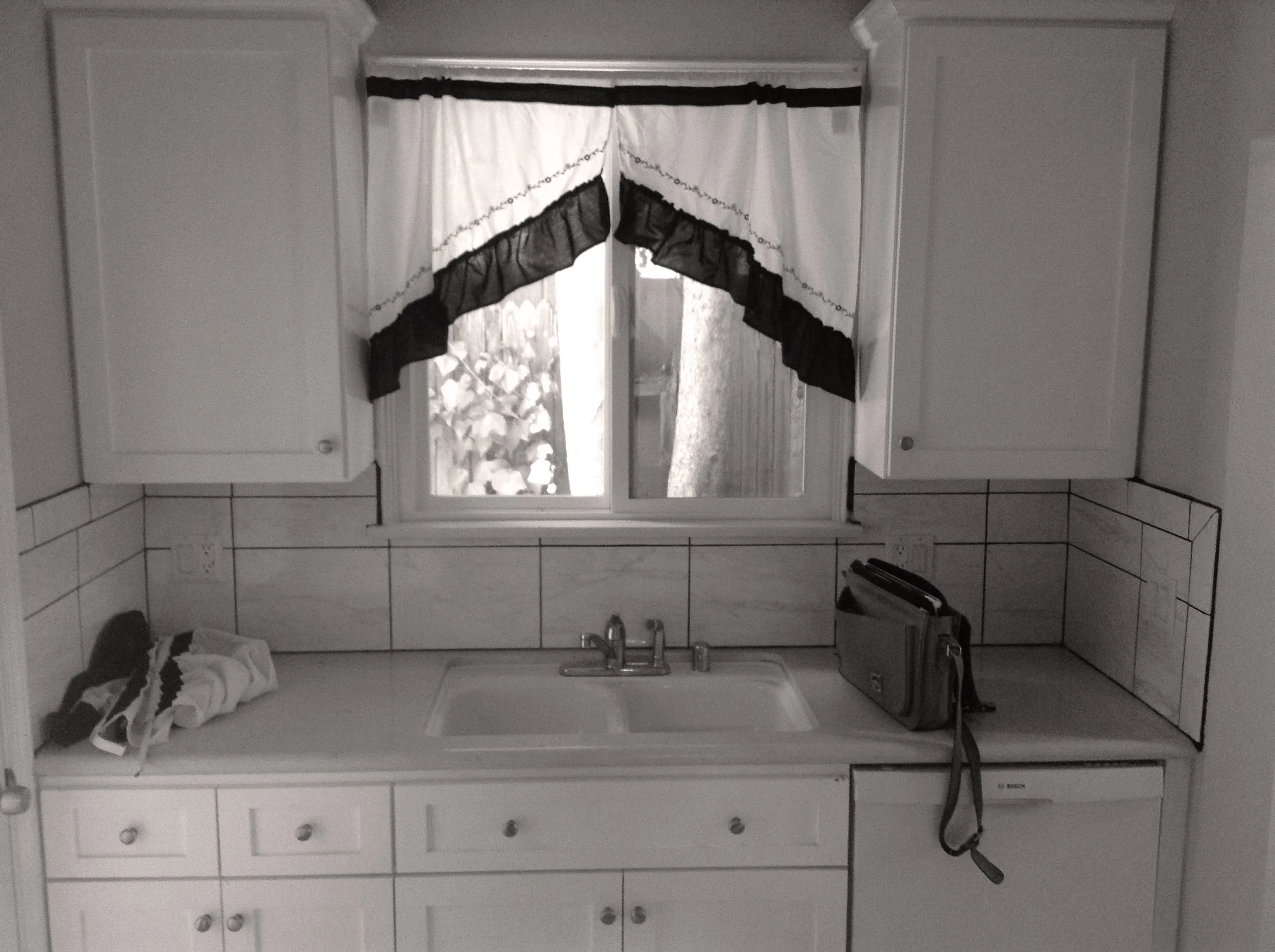 Black-and-white photo of a kitchen with a sink, window with curtains, cabinets, and a countertop with a small bag and clothes on it.