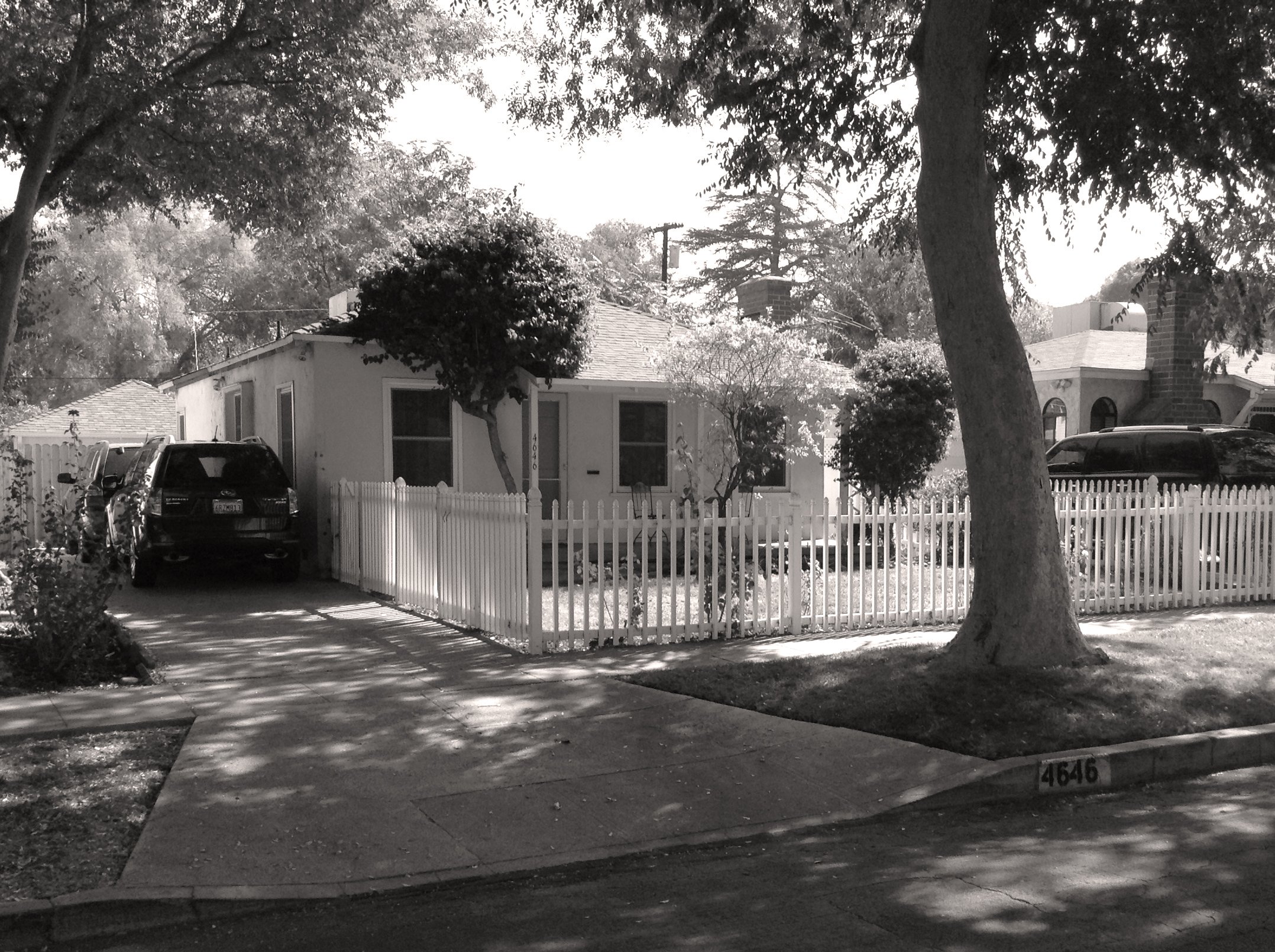 A small house with a white picket fence, trees providing shade, and cars parked in the driveway, in a residential neighborhood.