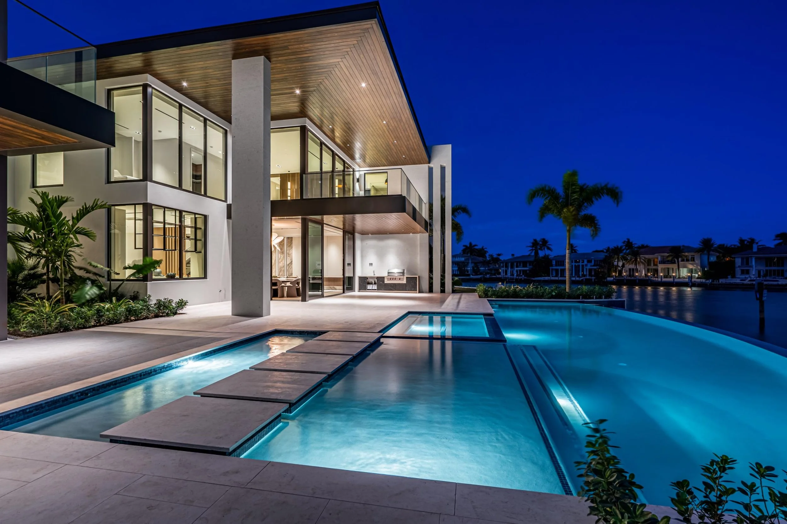 Modern house with large glass windows and a swimming pool at night, with palm trees in the background.