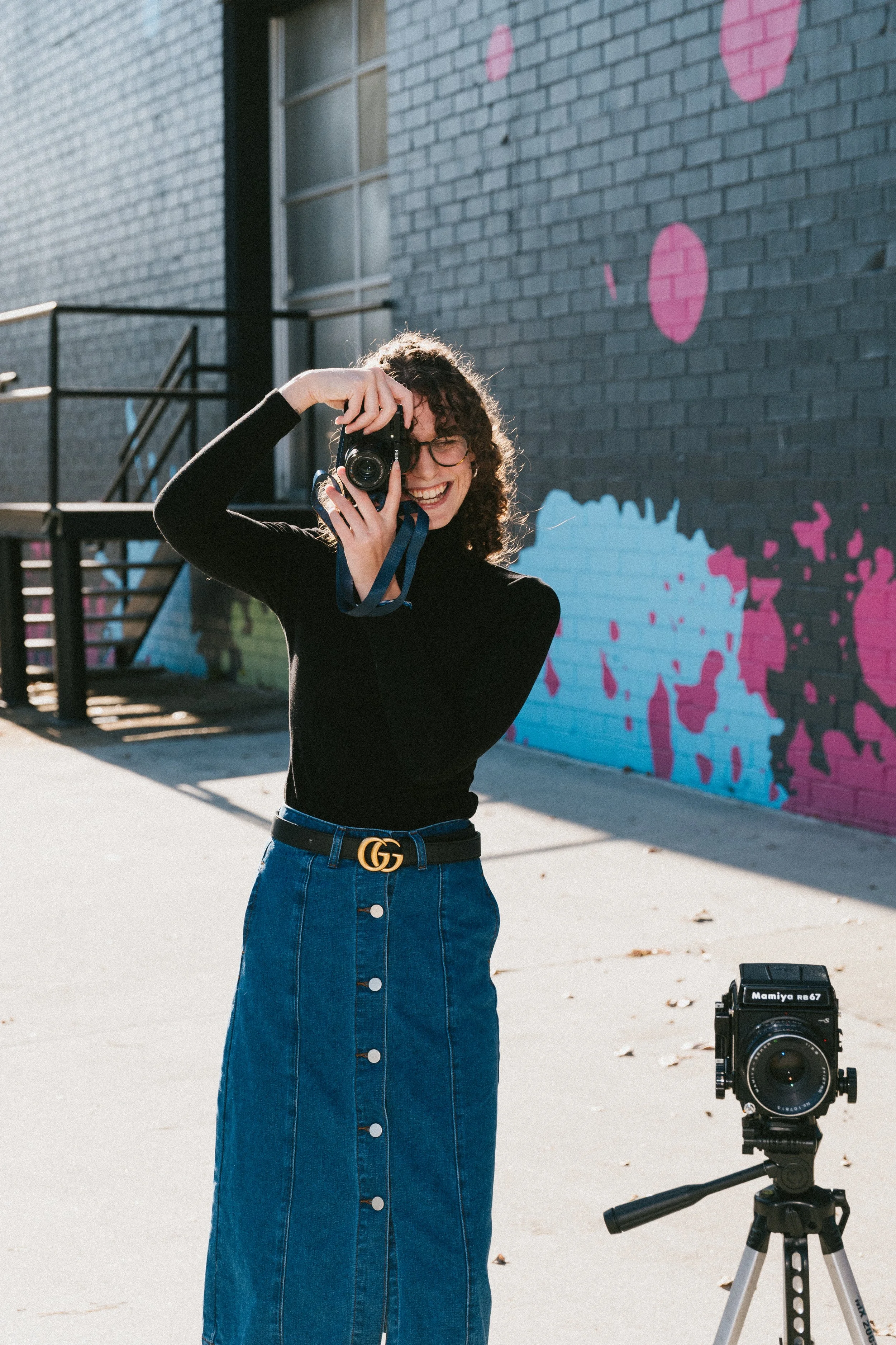 A woman with curly hair and glasses smiling and holding a camera. She is standing outdoors next to a colorful graffiti wall, with a professional camera on a tripod nearby.