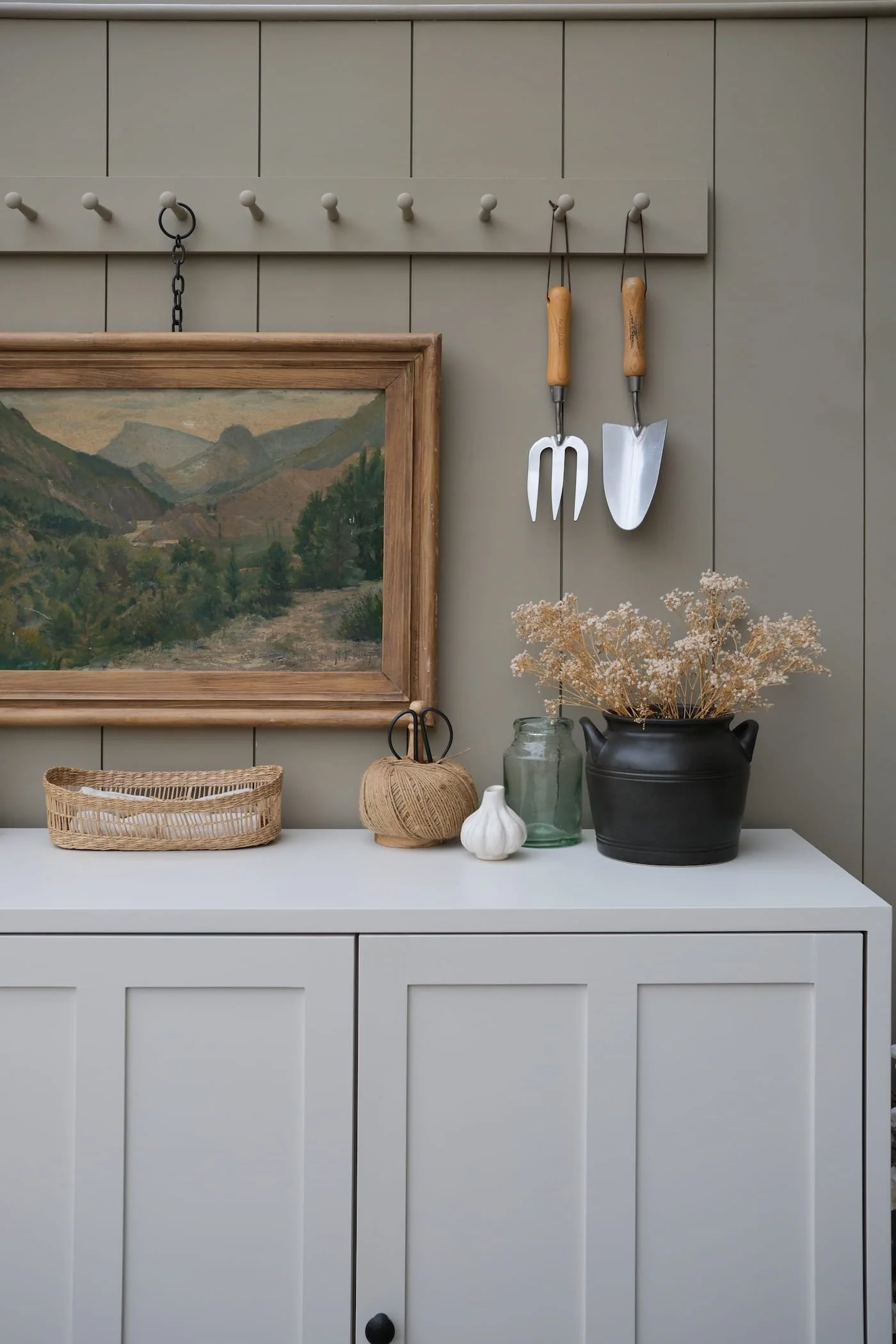 Boot room styling in a Gloucestershire cottage featuring vintage landscape artwork, hanging garden tools, wicker basket, ceramic accessories, and dried flowers – by Amy Broad Interior Design.