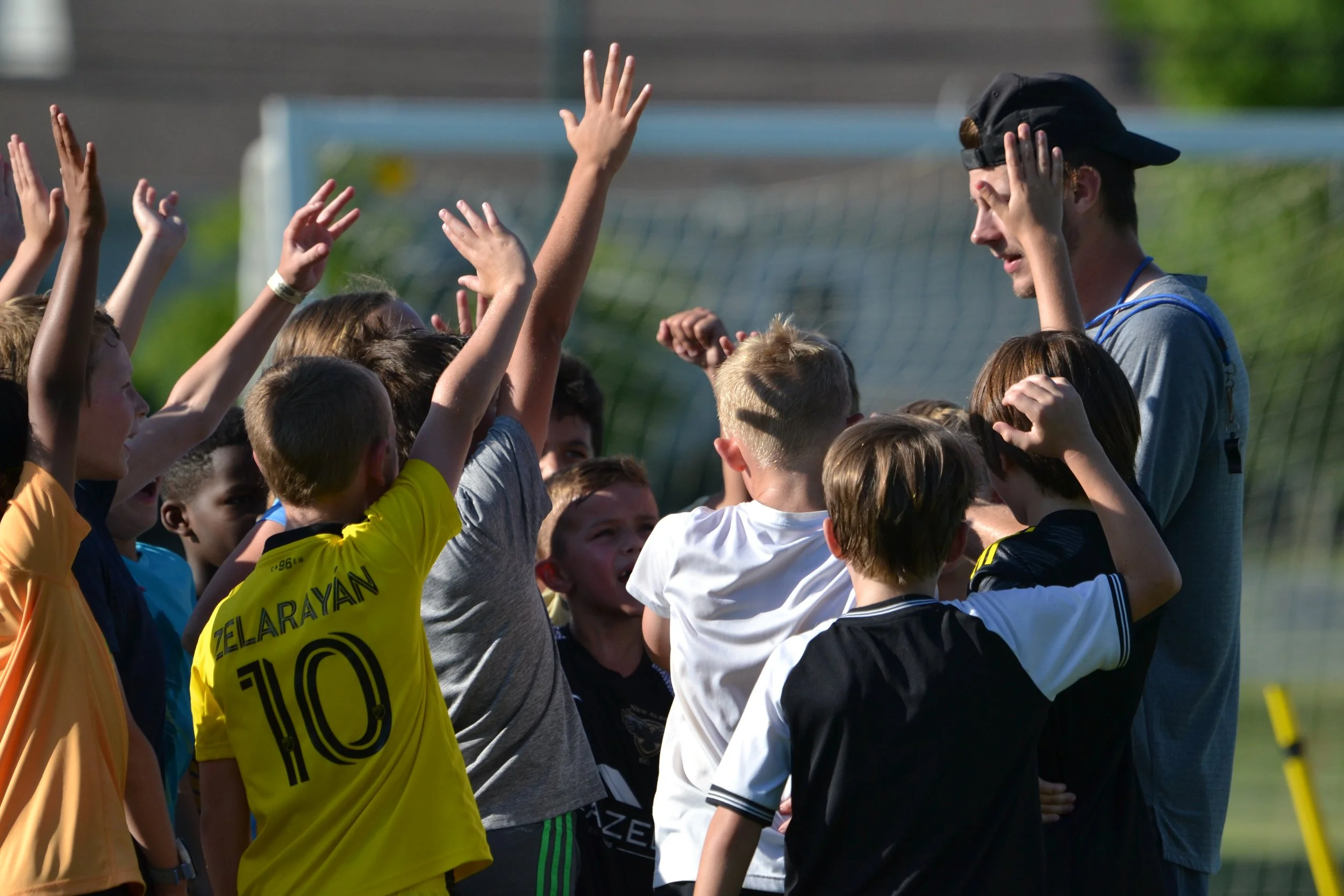 Children gather around a coach on a soccer field, raising their hands for a cheer or high-five, with a soccer goal in the background.