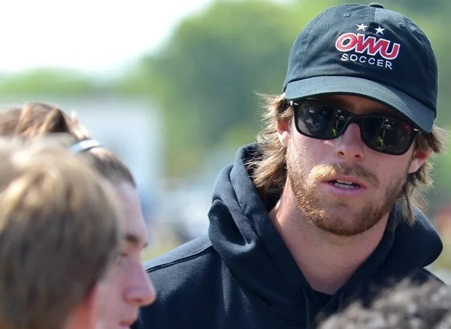 Man with long hair and beard wearing sunglasses, a black cap with 'OWU Soccer' logo, and a black hoodie, speaking to people outdoors on a sunny day.