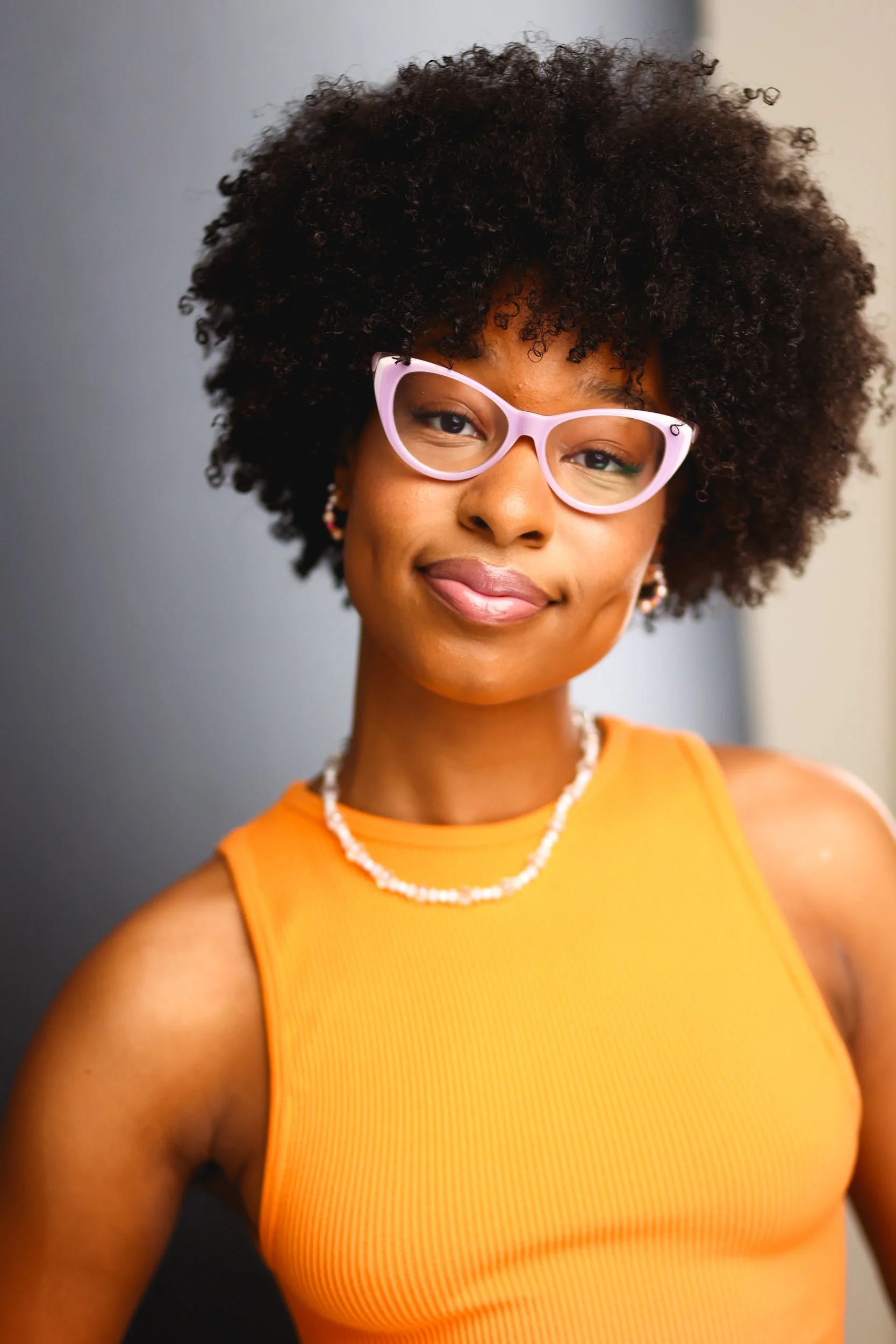 A woman with curly hair wearing pink glasses, an orange sleeveless top, a pearl necklace, and earrings, smiling at the camera against a neutral background.
