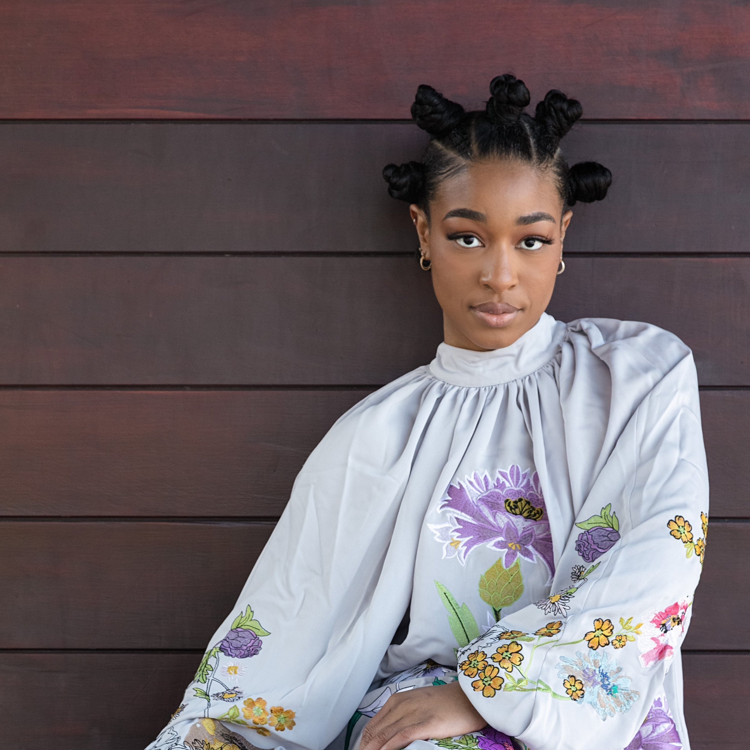 A young woman with styled bantu knots in her hair and hoop earrings, wearing a white top with colorful floral embroidery, sitting against a dark wooden wall.