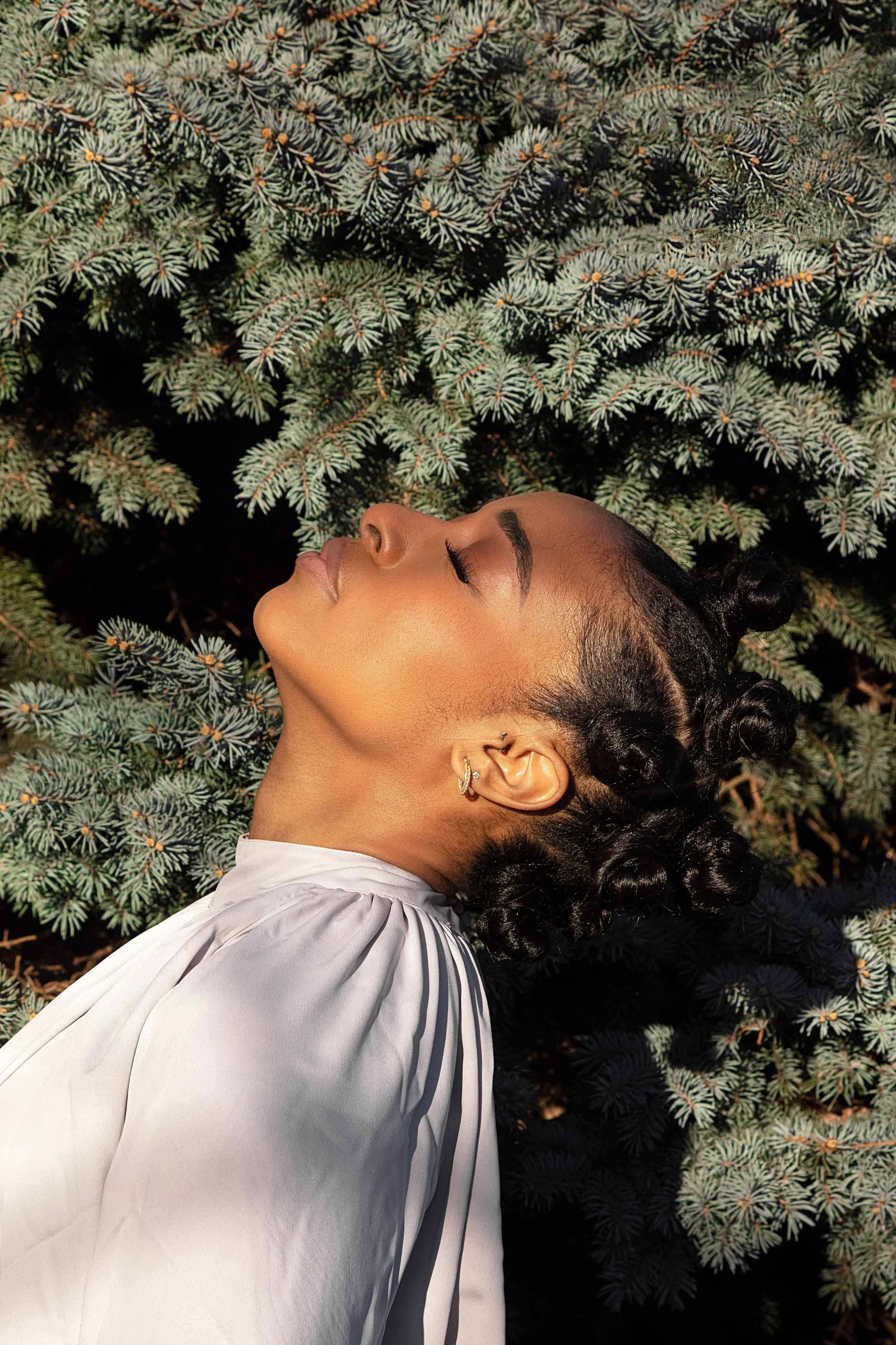 A woman with dark, styled hair lies on her back with her eyes closed, surrounded by green pine branches.