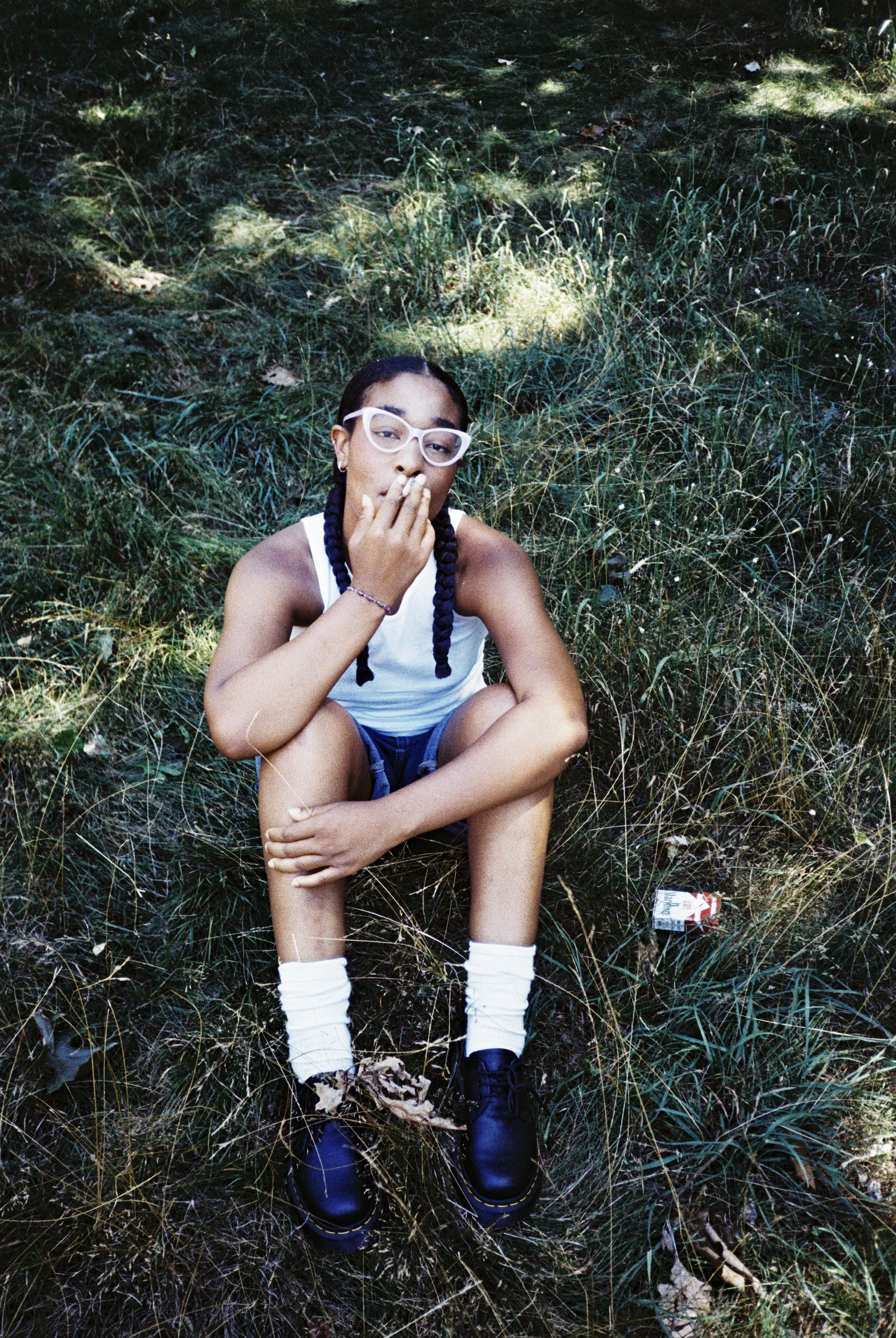Young person sitting on grass in a wooded area, wearing white glasses, a white tank top, navy shorts, white socks, and black shoes, with one hand near mouth and the other on knee.