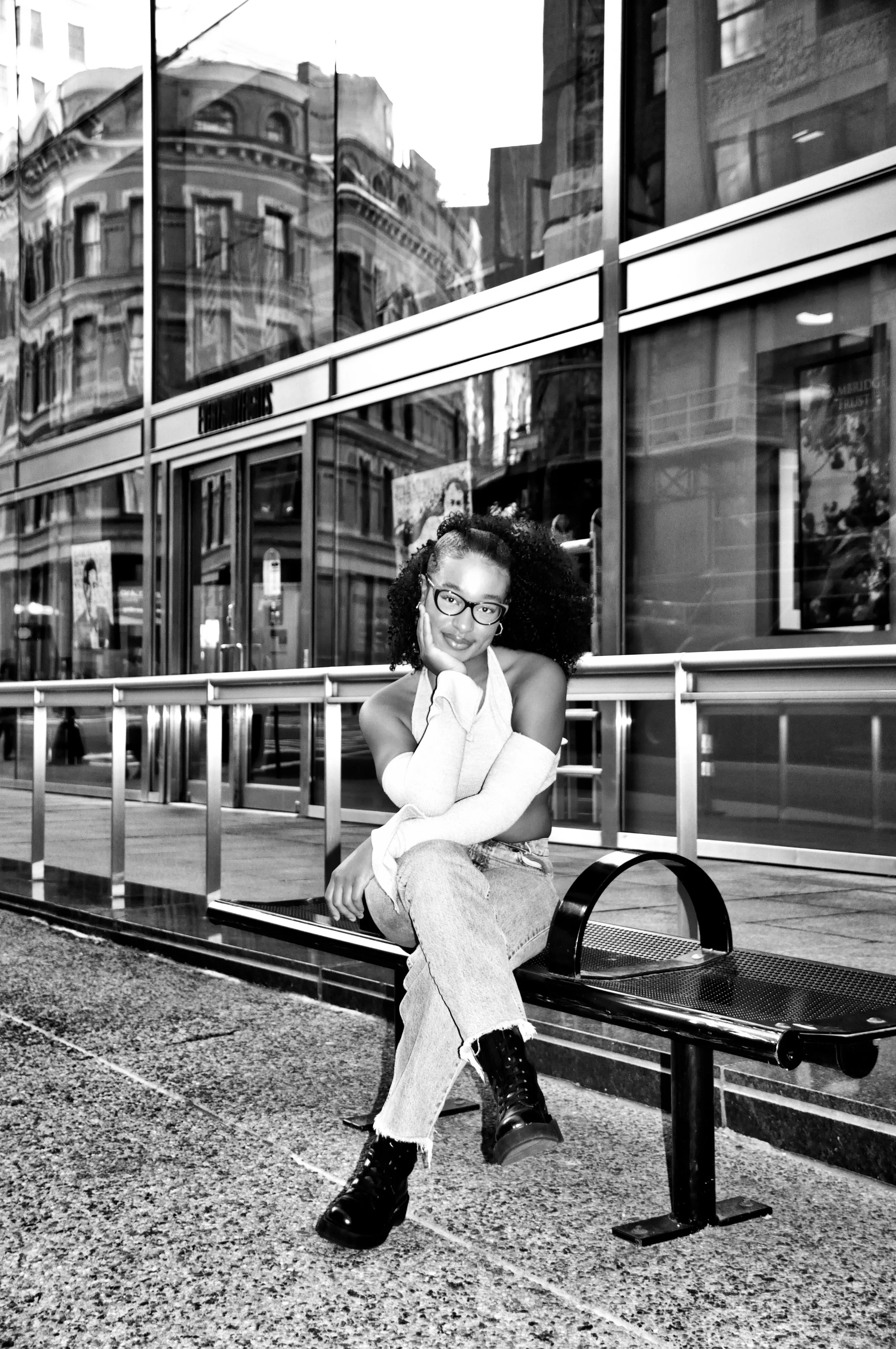 Black and white photo of a woman with curly hair and glasses sitting on a city bus stop bench, wearing ripped jeans, a long sleeve top, and boots, with a glass building and reflections of city buildings in the background.