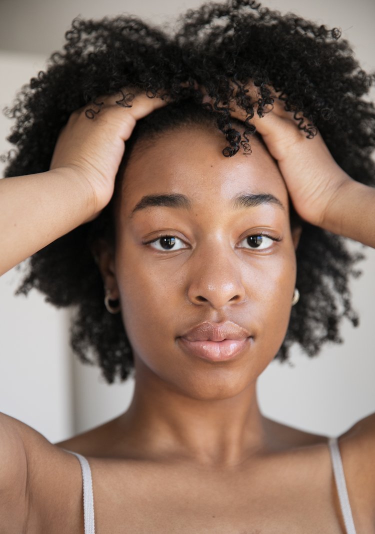 Close-up portrait of a young Black woman with natural curly hair, gently holding her head with both hands, looking directly at the camera with a neutral expression.