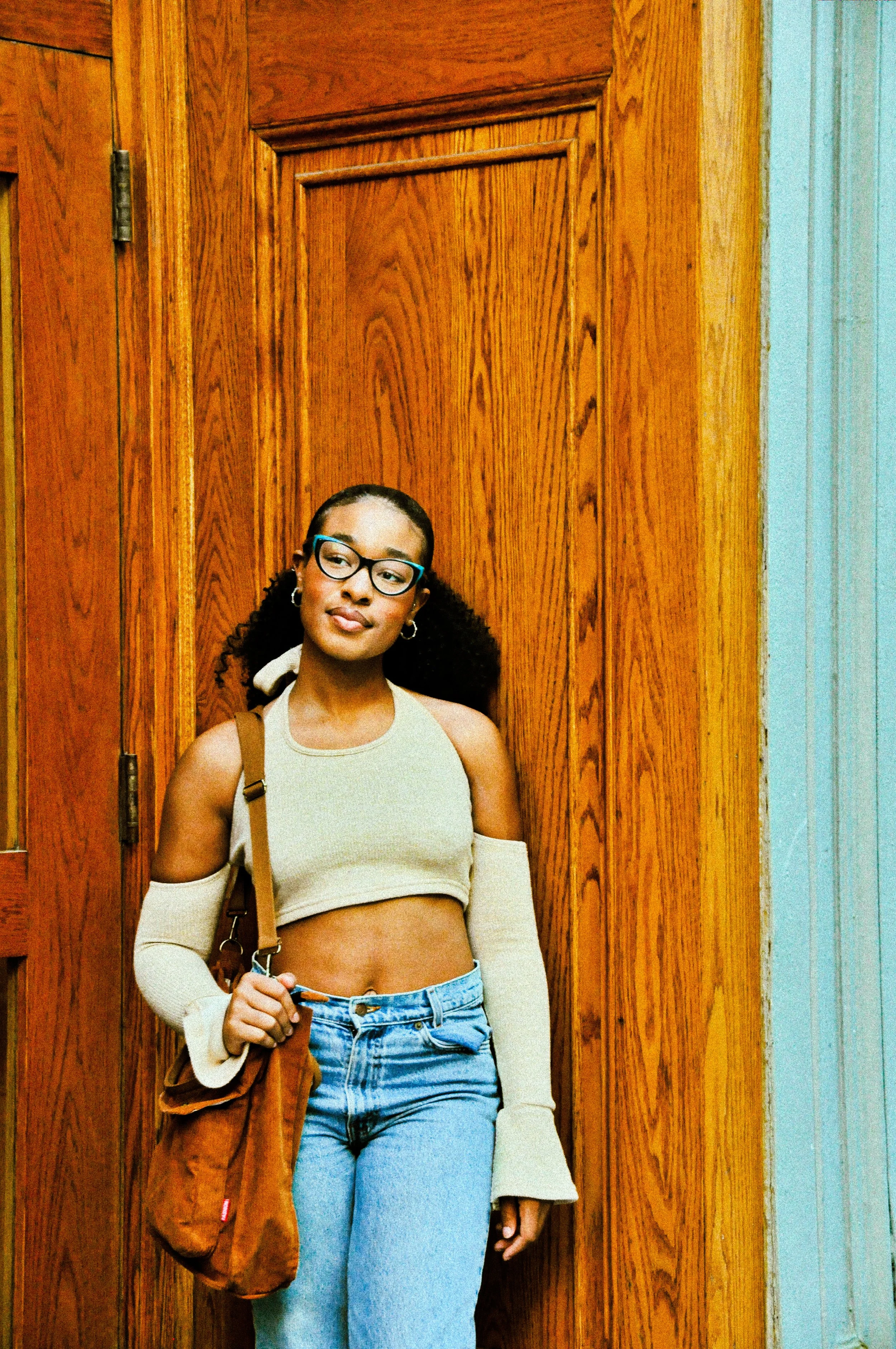 Young woman with glasses, curly hair, wearing a beige crop top with cut-outs on the shoulders, blue jeans, and carrying a brown bag, standing indoors against a background of wooden panels.