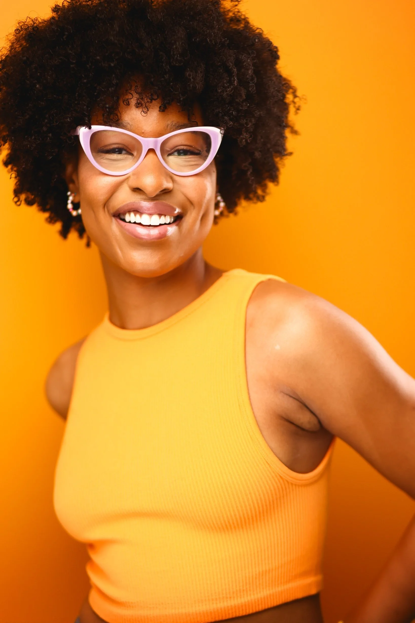 A smiling woman with curly hair wearing pink glasses and a sleeveless yellow top standing against an orange background.