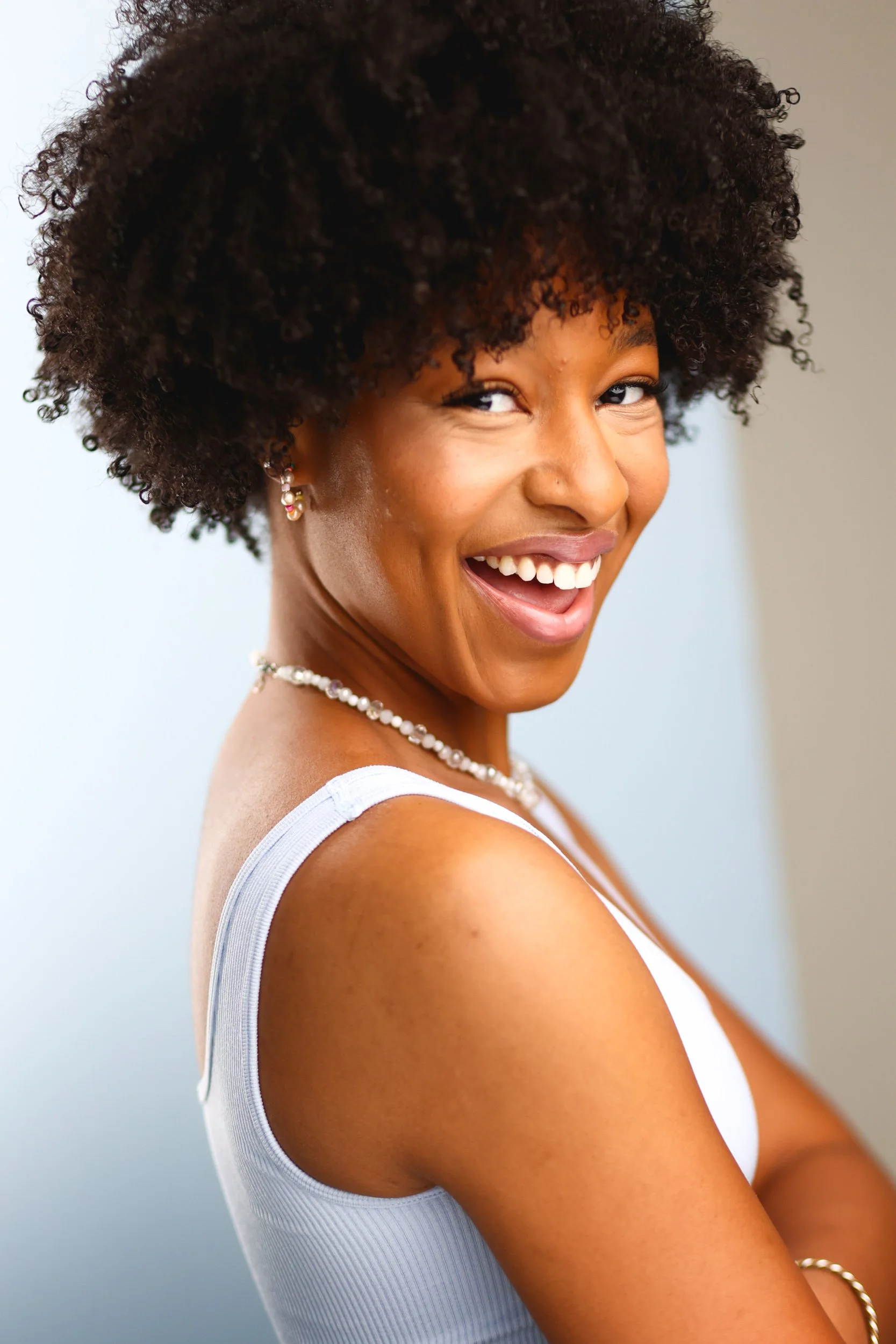 A woman with curly hair smiling and looking to the side, wearing a white sleeveless top and pearl jewelry.