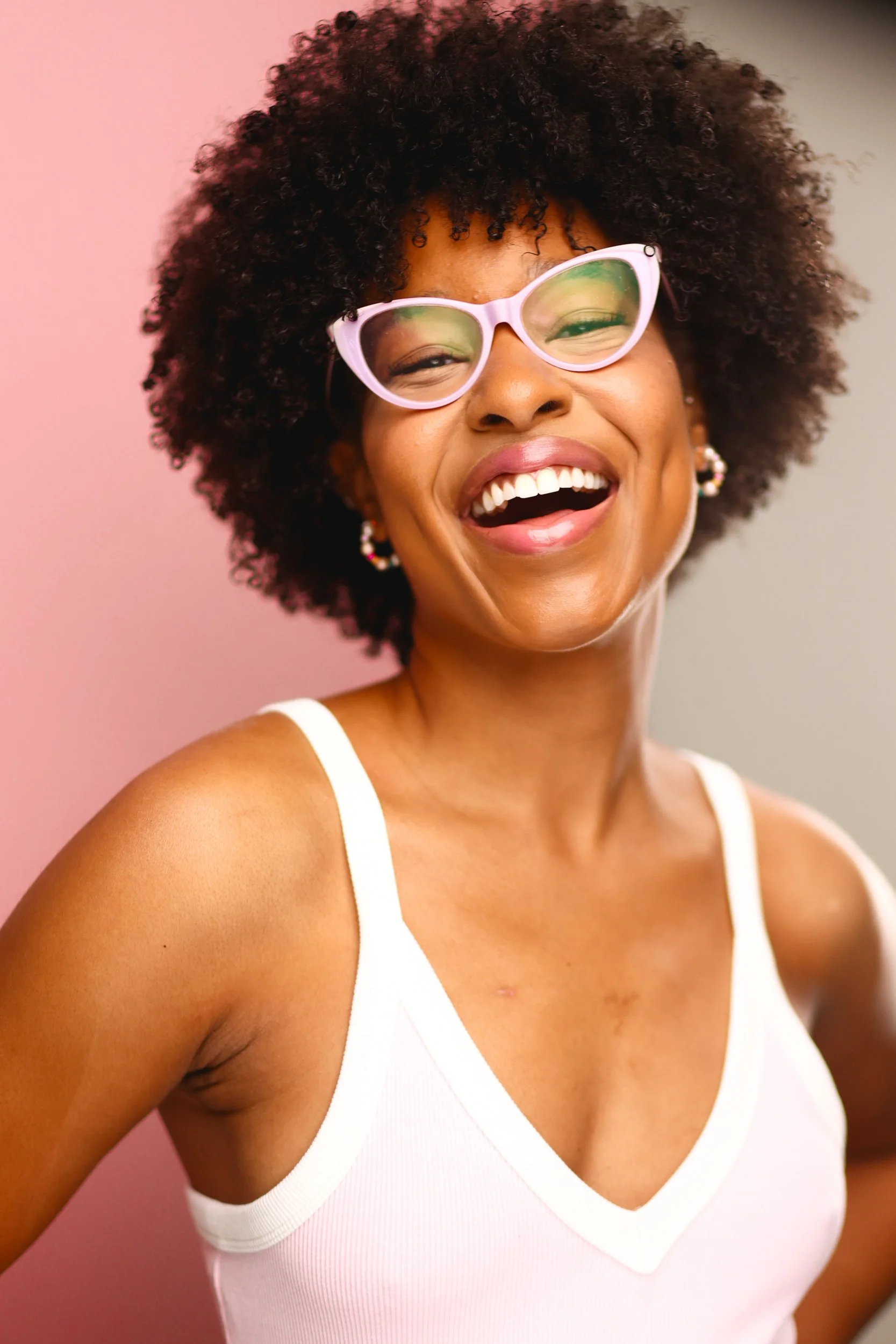 Smiling woman with curly hair wearing pink glasses and a white tank top, standing against a pink and gray background.