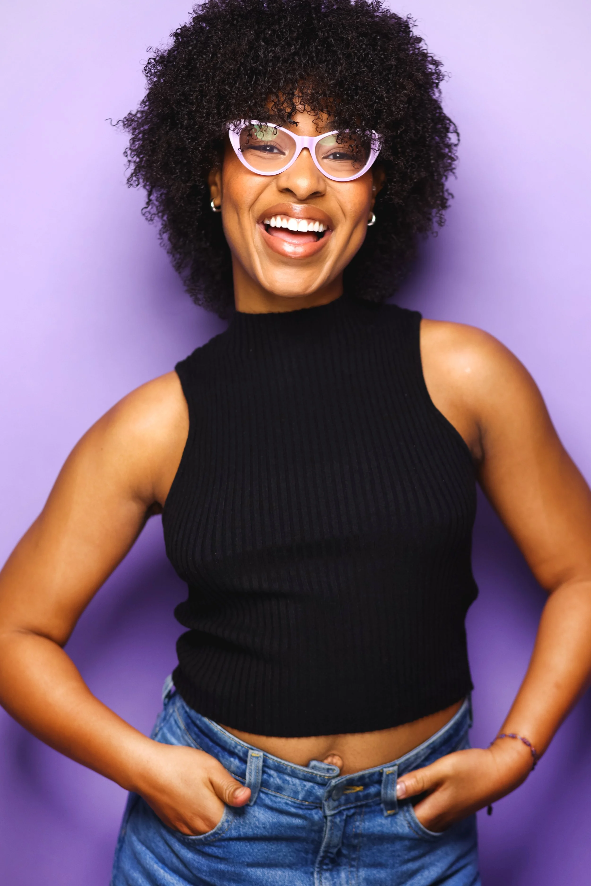 A woman with curly hair, wearing pink eyeglasses, a black sleeveless top, and blue jeans, smiling and holding her hands in her pockets against a purple background.
