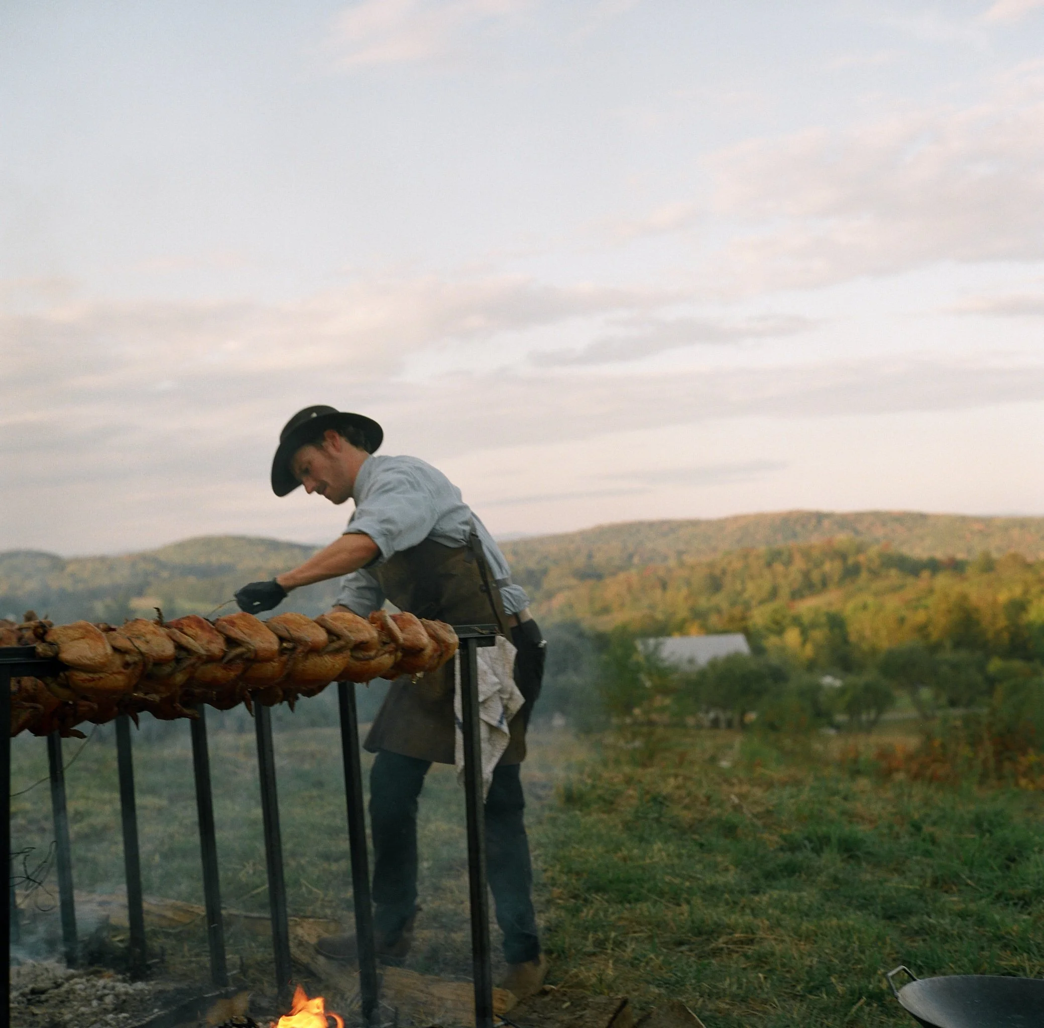 A man wearing a cowboy hat and apron is grilling multiple chickens on a horizontal rotisserie outdoors during sunset, with rolling hills and trees in the background.